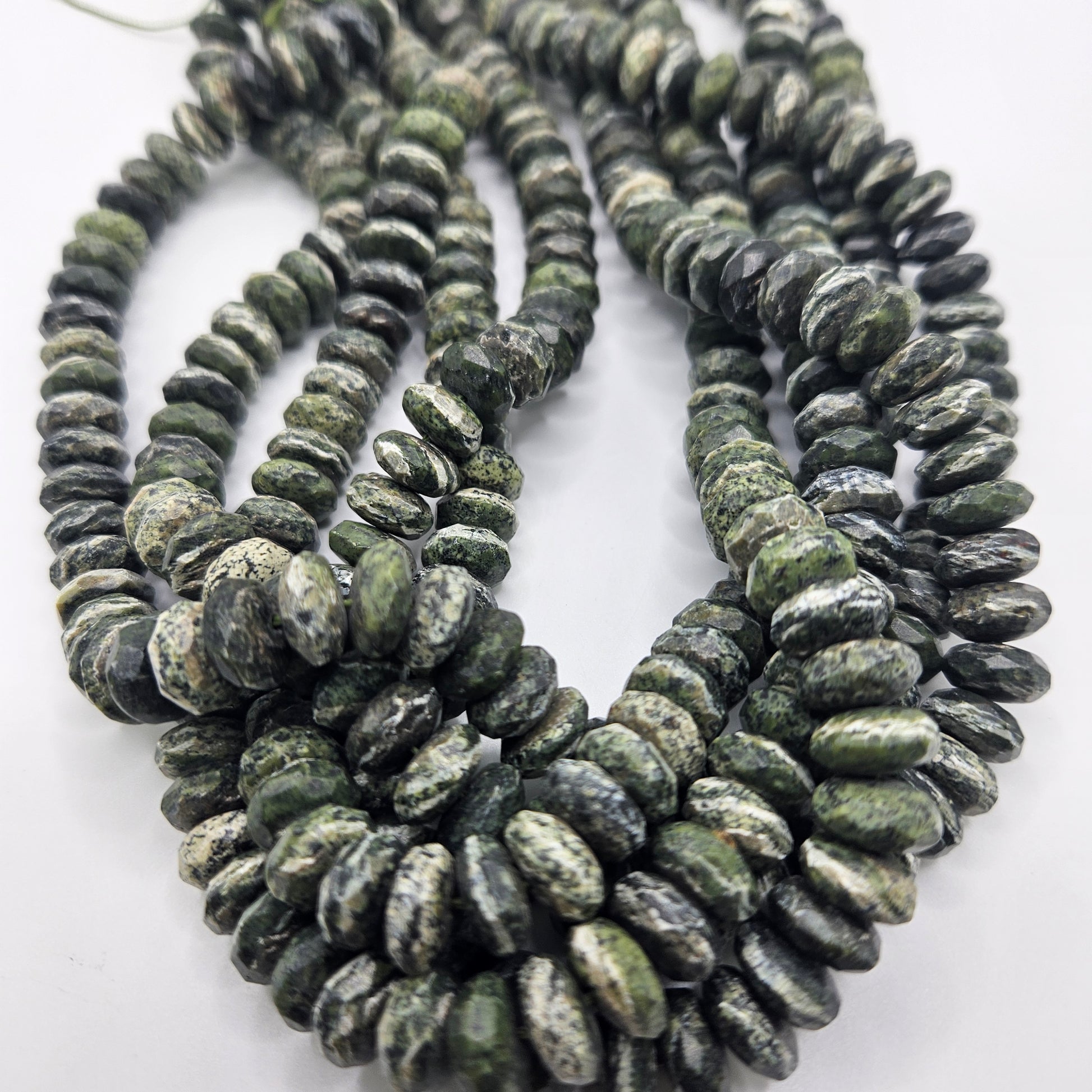 A close-up view highlighting multiple strands of the 10mm Green Jade Rondelle Bead Strand, along with gray marbled stone beads, emphasizes their smooth, rounded shapes and intricate patterns. These strands are artfully arranged in overlapping loops on a white surface.