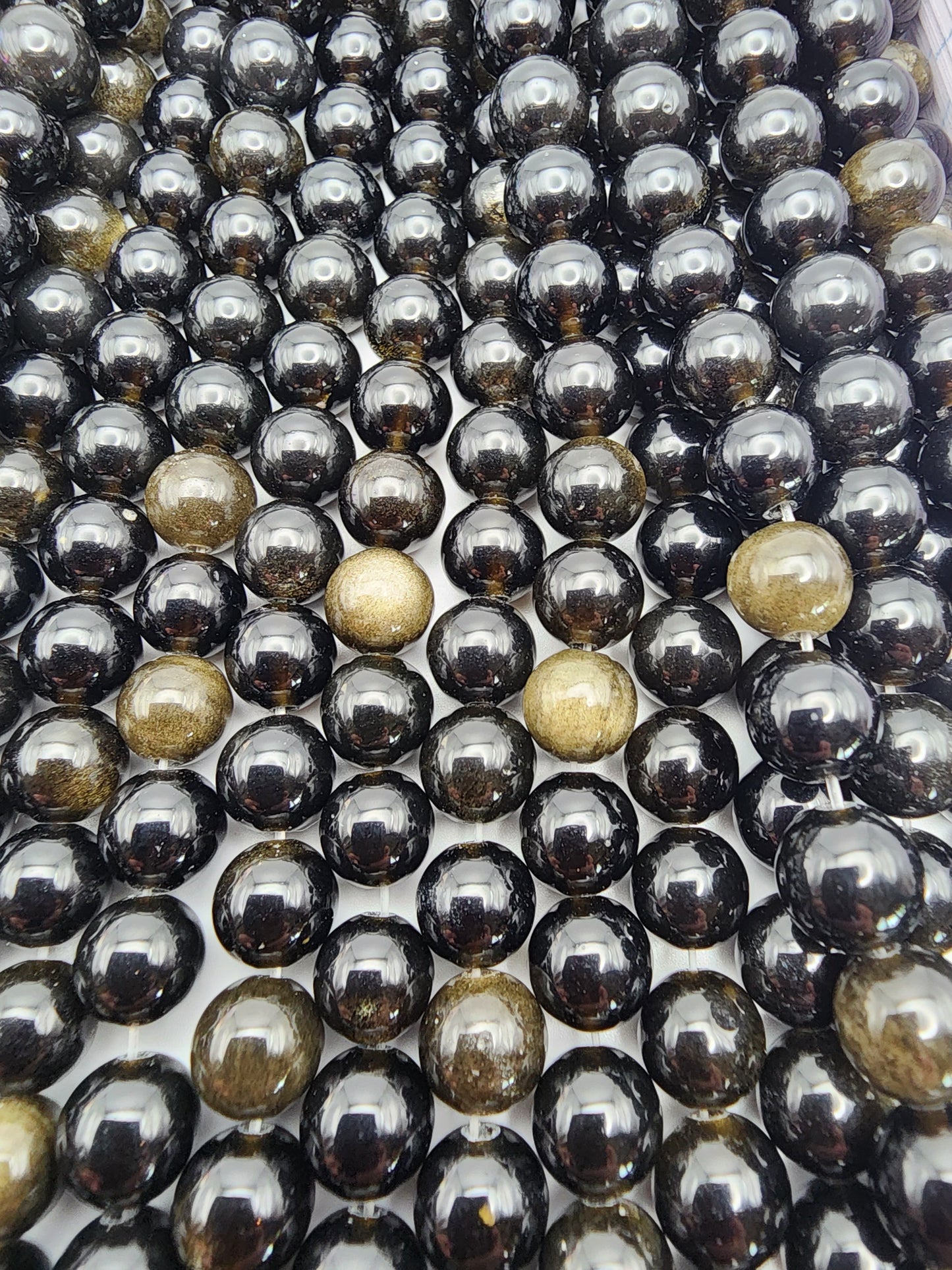 Rows of shiny 8mm Gold Obsidian Rounds with a few gold marbles interspersed throughout create a visually striking pattern on a white surface.