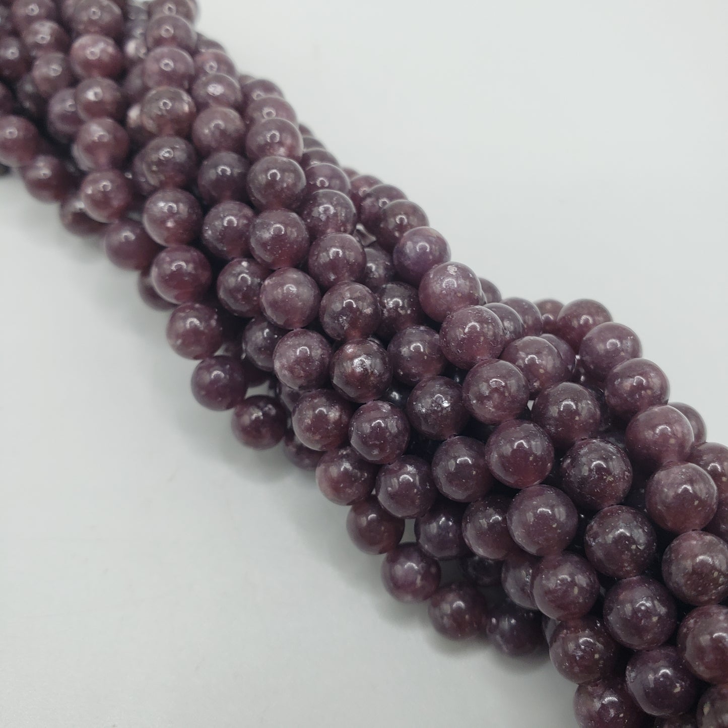 Close-up of several strands of 4mm Lepidolite beads, showcasing their dark purple hue and glossy finish as they are arranged in a twisted pattern on a white surface, subtly reflecting light.