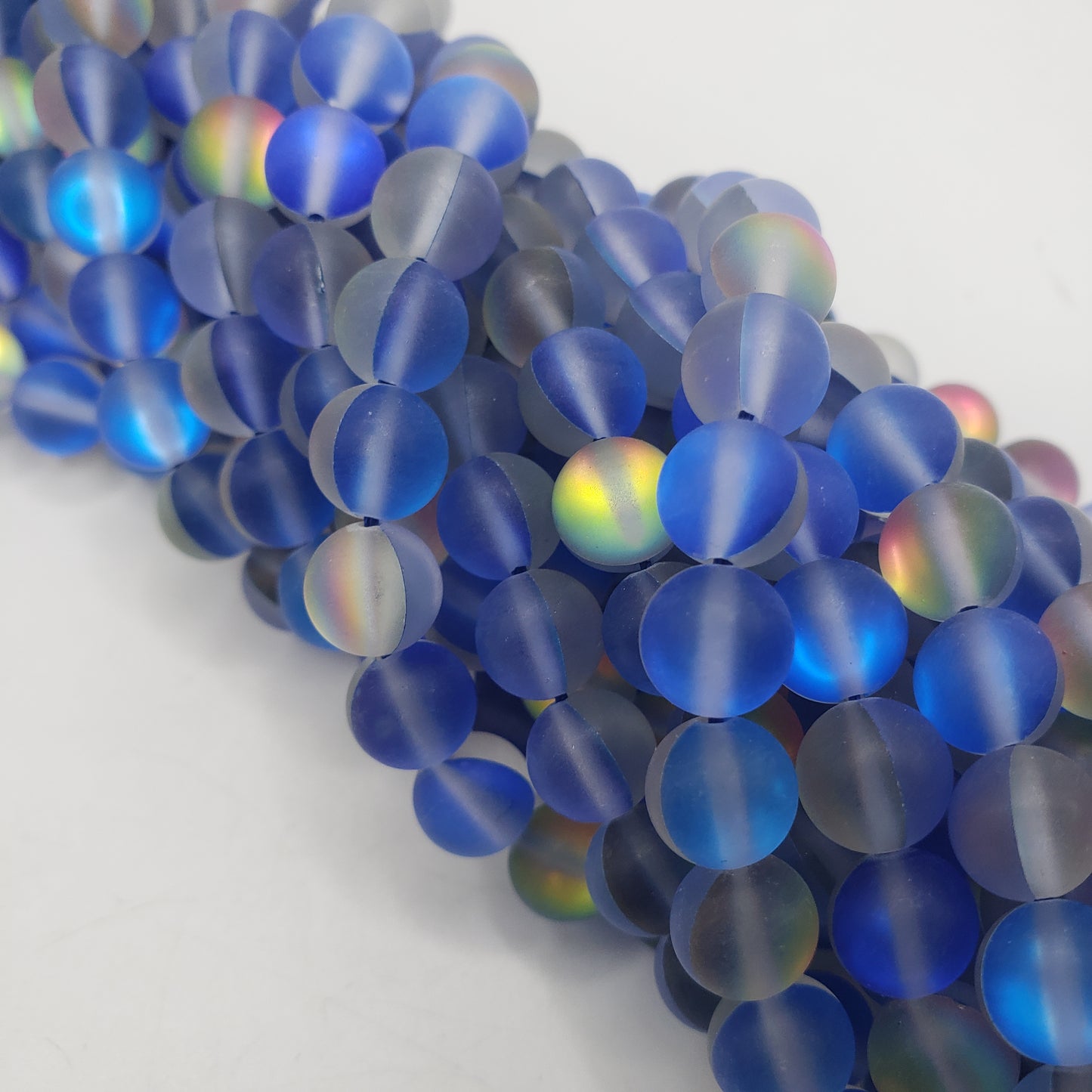 Close-up of a cluster of 8mm Blue Matte Mermaid Glass beads, showcasing their iridescent qualities with subtle rainbow hues under light. These beads form a vibrant and colorful pattern against a light gray background.