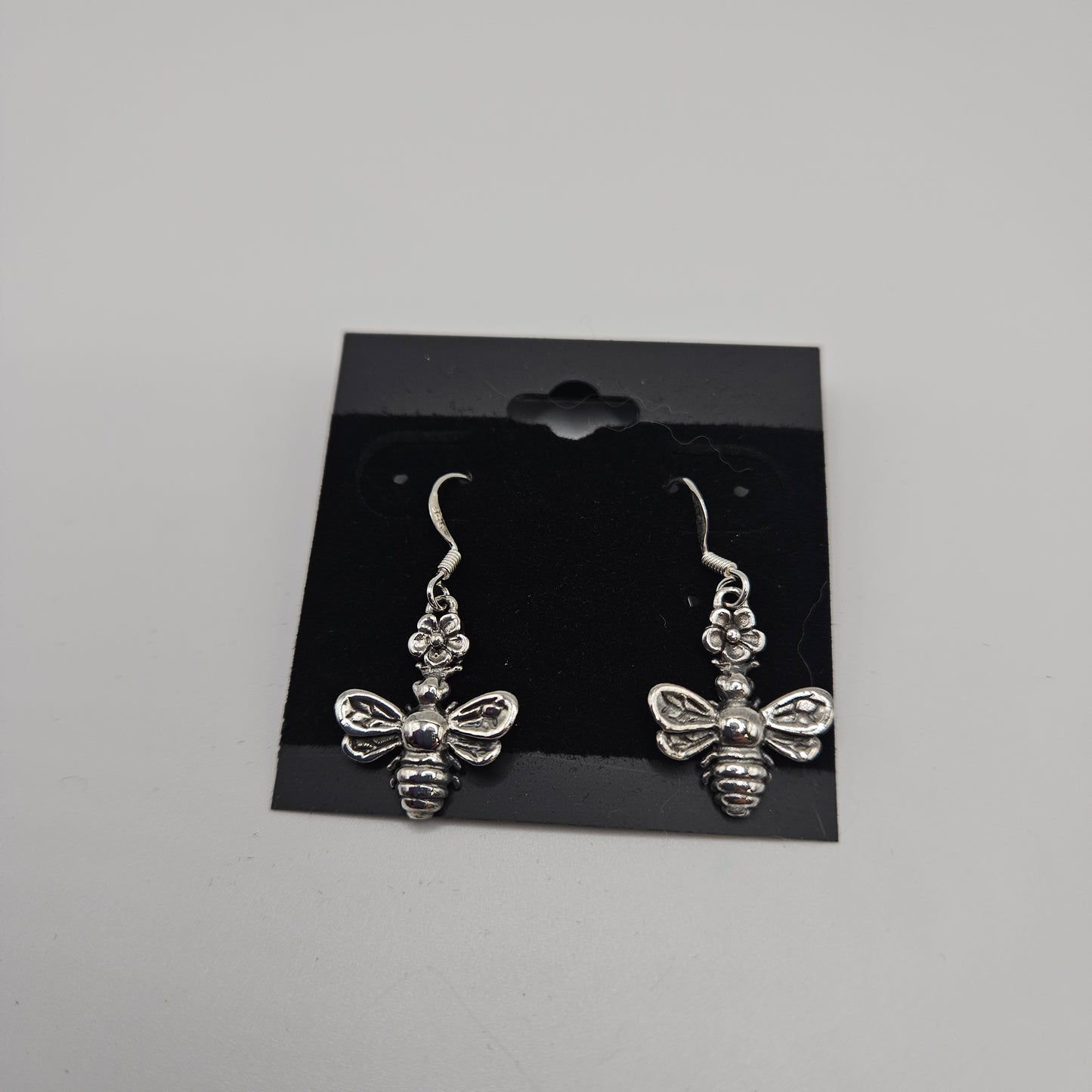 Introducing the SS Bee Earrings, priced at $33.99, featuring a pair of silver earrings intricately designed in the shape of bees with detailed wings and bodies. These stunning earrings are beautifully presented on a black card equipped with hooks for easy display against a neutral gray backdrop.