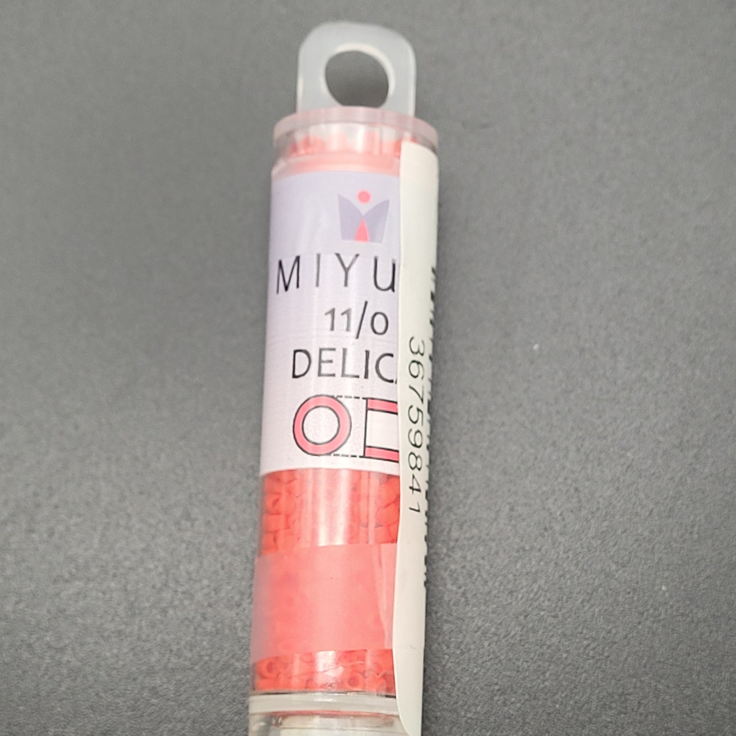 A small, clear plastic tube holding matte opaque light siam red beads with the label "11/0 DELICA BD MATTE OPAQUE LT.SIAM APRX 7.2 GM DB757-TB" is placed on a gray surface.