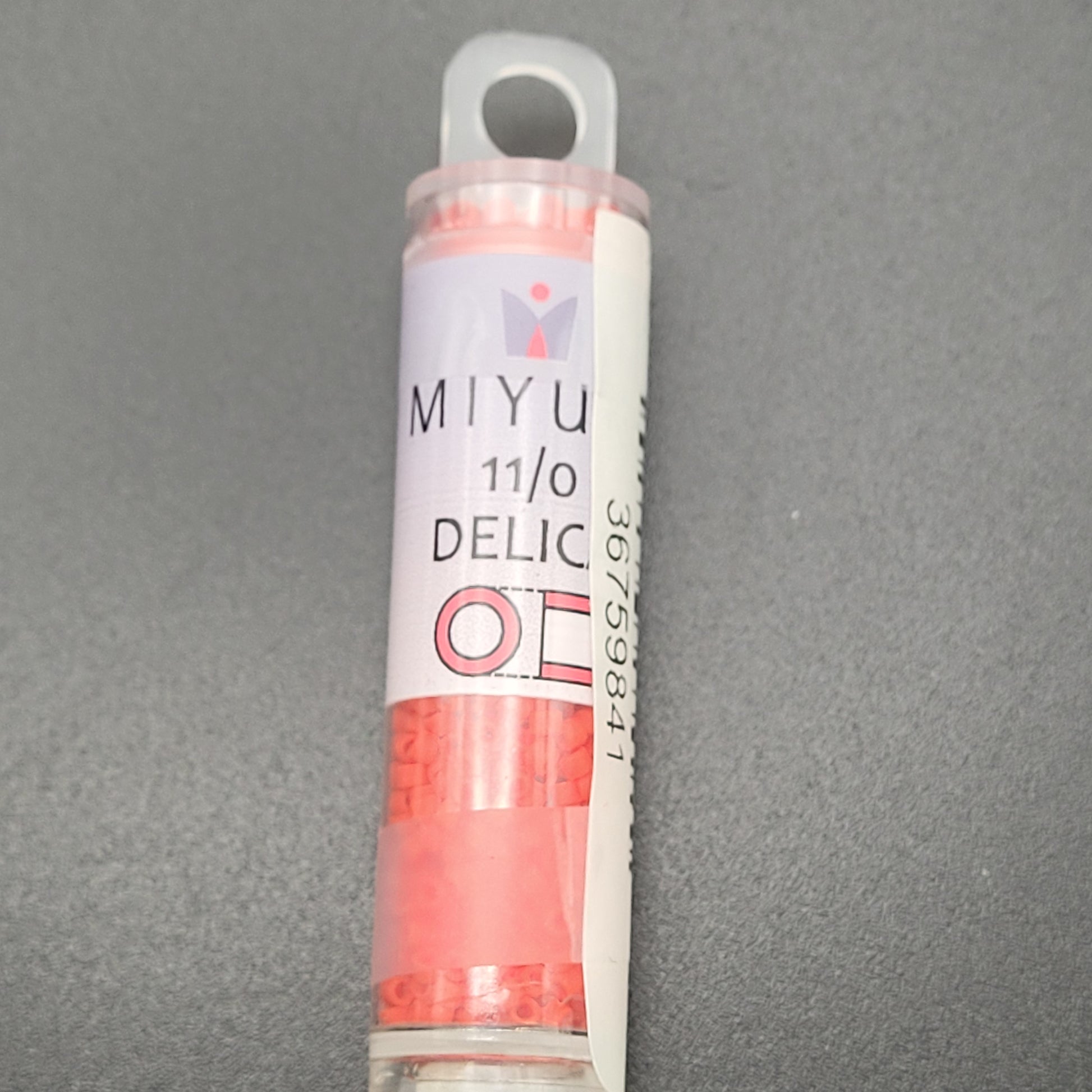 A small, clear plastic tube holding matte opaque light siam red beads with the label "11/0 DELICA BD MATTE OPAQUE LT.SIAM APRX 7.2 GM DB757-TB" is placed on a gray surface.