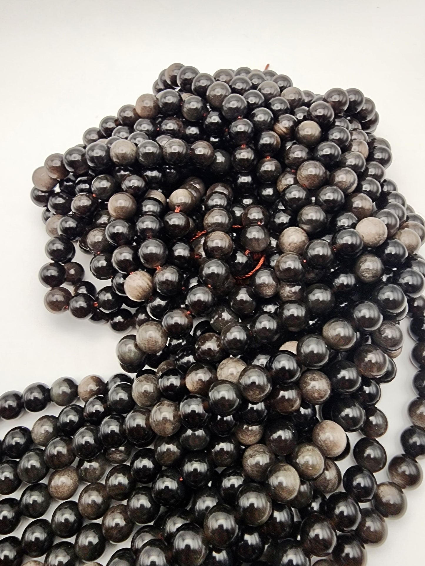 A collection of 8mm Silver Obsidian Round beads is loosely arranged on a white surface. These beads are dark, shiny, and round with a smooth texture that reflects light.