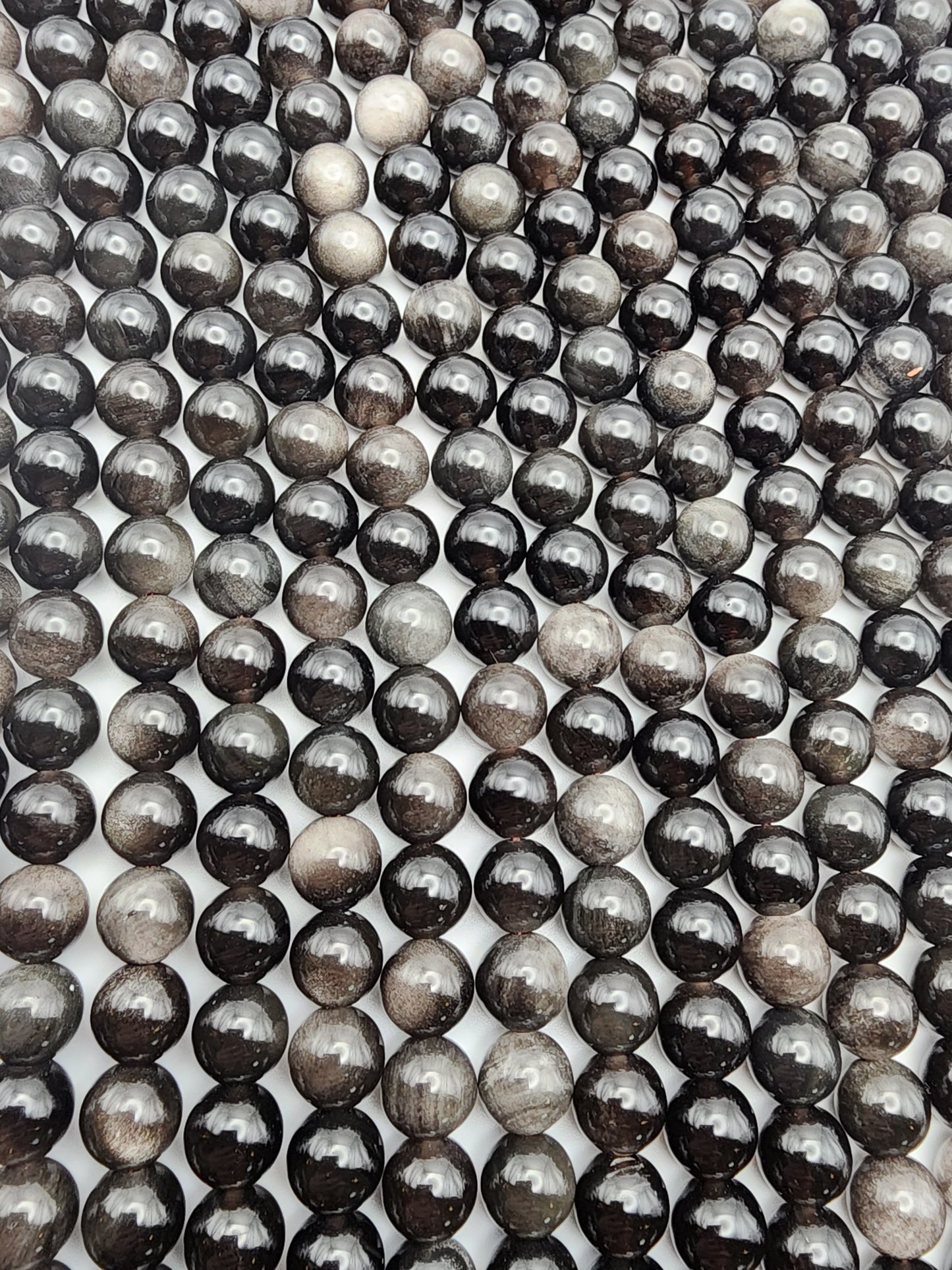 Rows of glossy 8mm Silver Obsidian Round beads, with a black and gray marble-like appearance, are arranged closely together. They create a captivating pattern of reflections and varying shades against a white backdrop.
