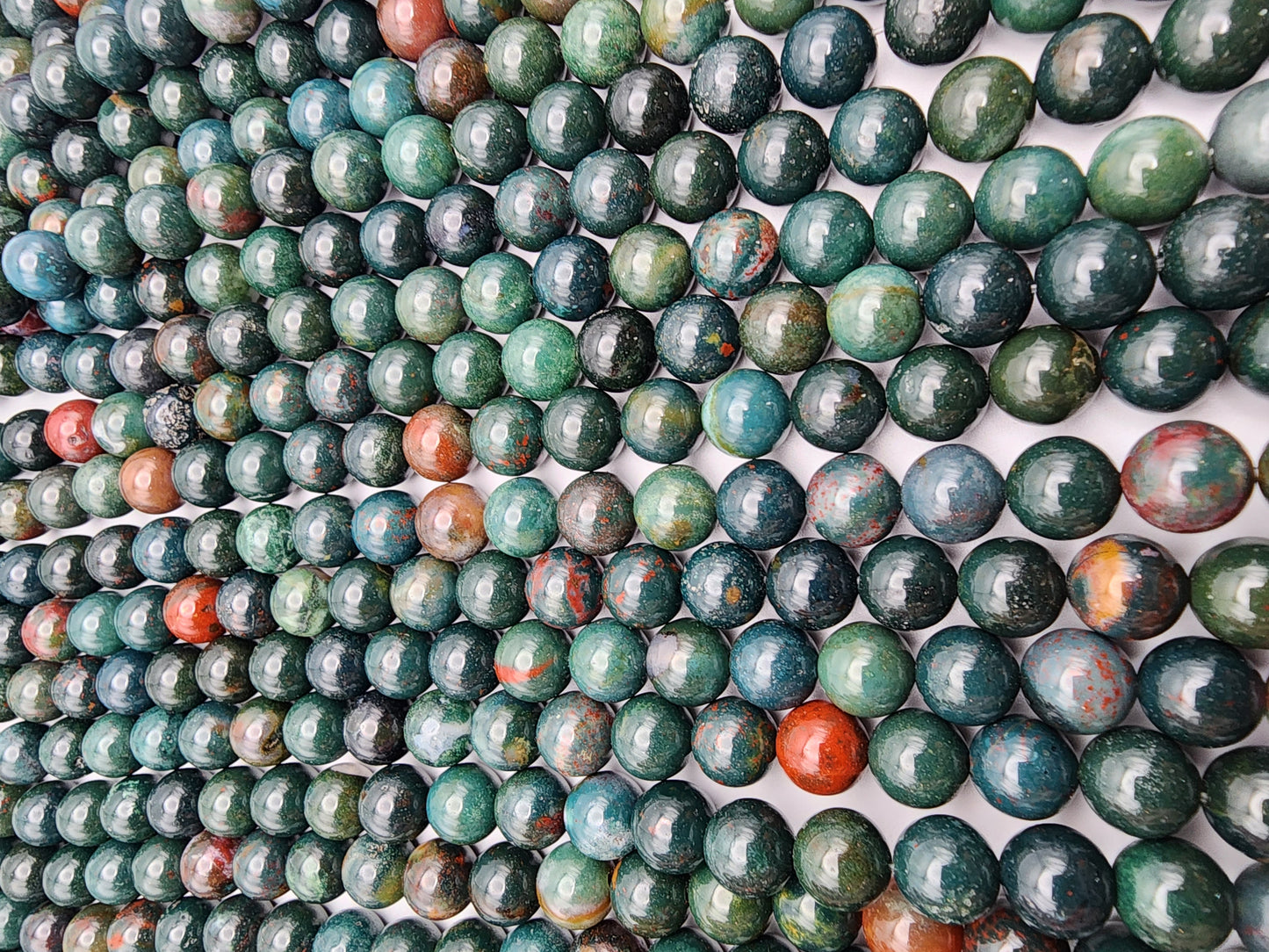 A close-up view of a collection of 8mm Bloodstone Rounds arranged in rows. These stones display a variety of colors, including shades of green, blue, and red, with swirling patterns. The reflective surface of each bloodstone catches the light.