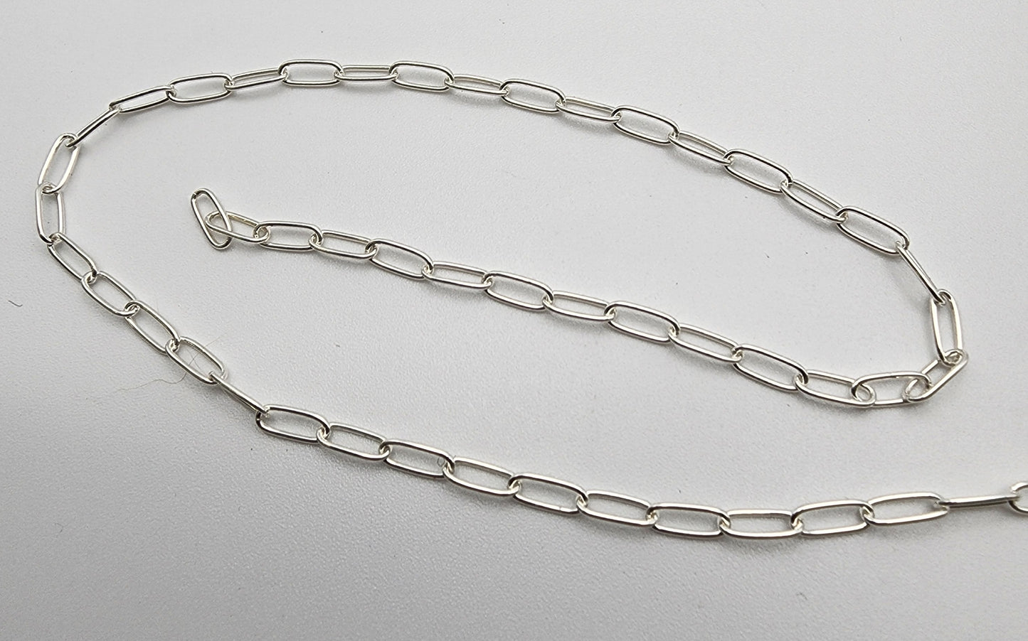 The SS Paper Clip Chain 2.5mm, a delicate silver chain with elongated oval links, is laid out on a white surface. The chain is partially coiled, showcasing its simple design and shine.