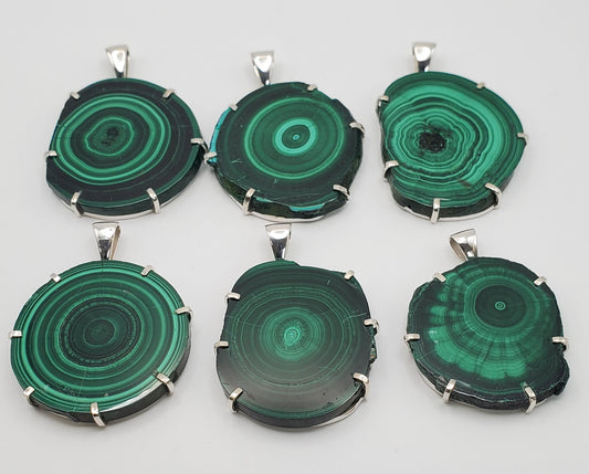 Six SS Malachite Large Stalactite Pendants with unique green concentric circle patterns are elegantly displayed on a white surface. Each pendant, encased in metal, features individual banded designs reminiscent of a handmade ring collection available for purchase online.