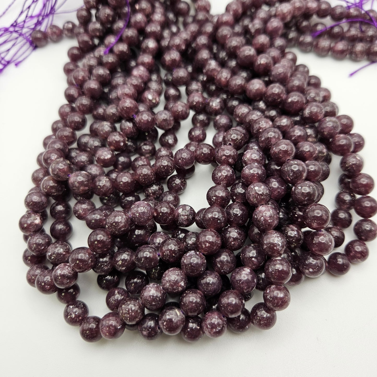 Close-up of several strands of 8mm Lepidolite round beads with speckles, arranged in loops on a white background. The beads have a shiny, polished surface, and the strands end with purple thread.
