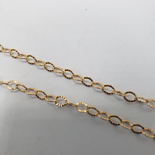 The GF Diamond Cut Oval Link Chain is shown against a plain white background, featuring two strands of delicate gold chain with oval links. The close-up view emphasizes the chain's intricate link pattern and glossy finish.