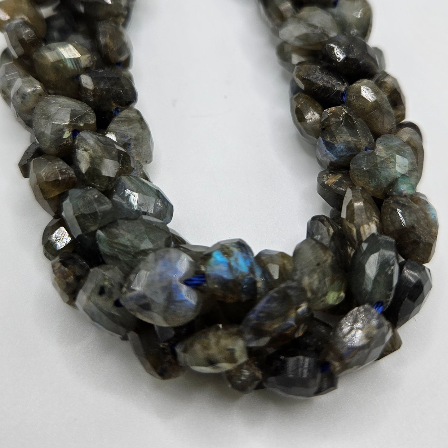 A close-up photograph showcases the 8mm Labradorite Faceted Heart Bead Strand. The irregularly shaped beads display grey tones with flashes of blue and green iridescence, characteristic of this unique gemstone. The strand is elegantly displayed on a plain white background.