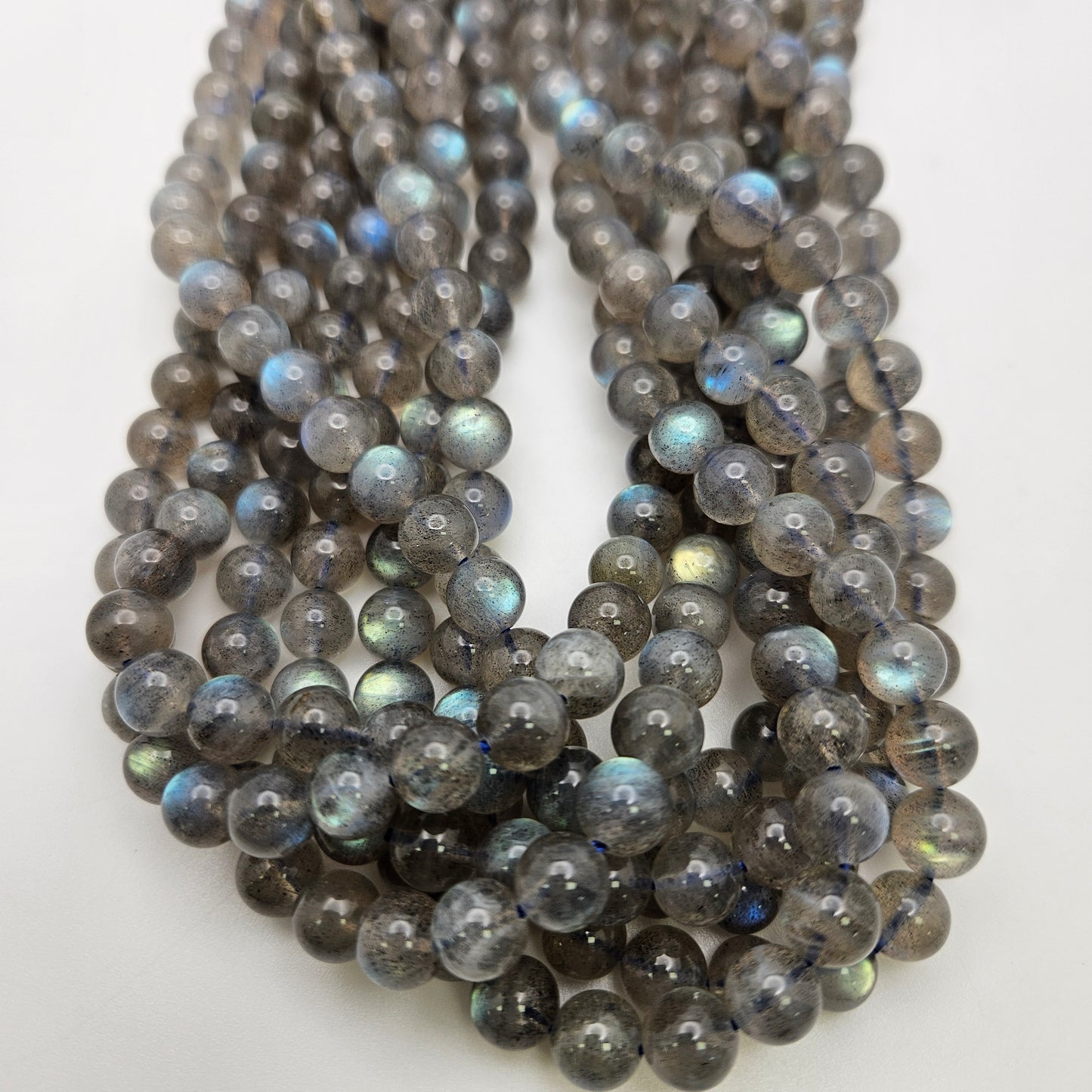 A collection of 8mm Labradorite Round beads featuring a shiny, iridescent surface that displays a mix of gray, blue, and green hues, closely arranged against a white background.