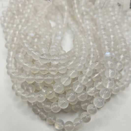 Close-up of several 8mm Blue Moonstone Round Bead Strands with a glossy finish, arranged on a surface. The translucent white beads appear uniform in size and are closely packed together.