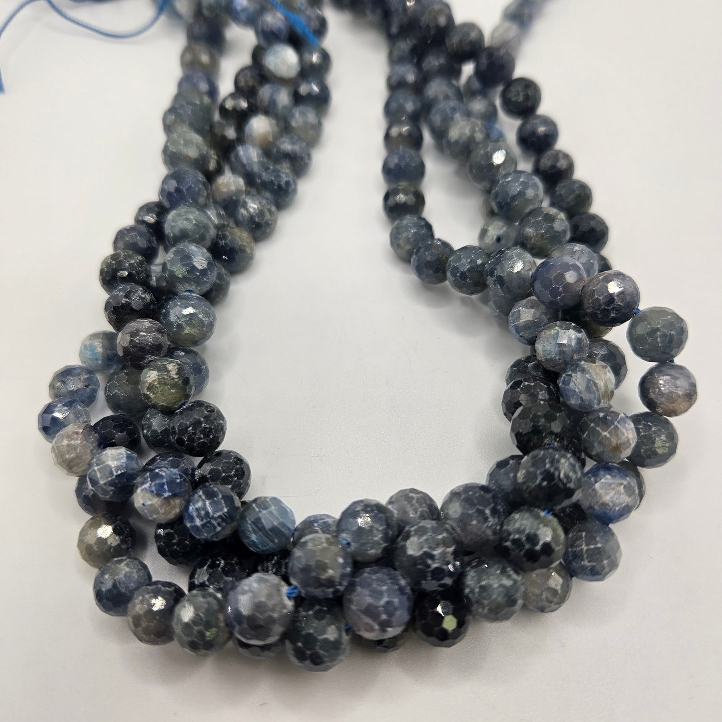 A close-up image showcases multiple strands of the 8mm Kyanite Faceted Round Bead Strand, featuring dark gray and blue hues. The slightly reflective beads are elegantly organized in loops against a plain white backdrop.