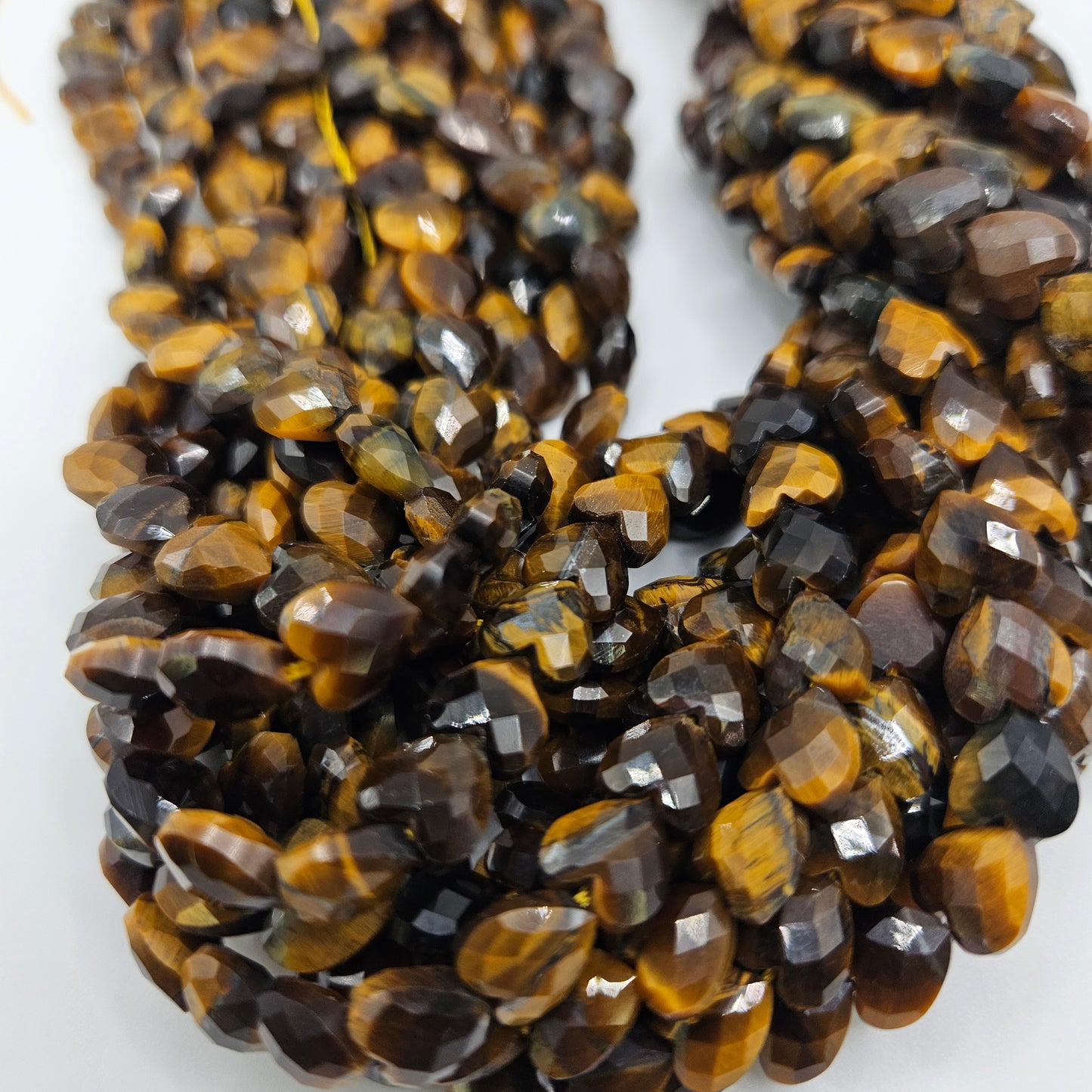 Close-up of multiple strands of 8mm Tiger's Eye Faceted Heart Beads. The polished beads boast a rich, smooth texture and showcase a blend of deep brown and golden reflective bands, creating a striking, shimmering effect. The strands are loosely coiled against a white background.