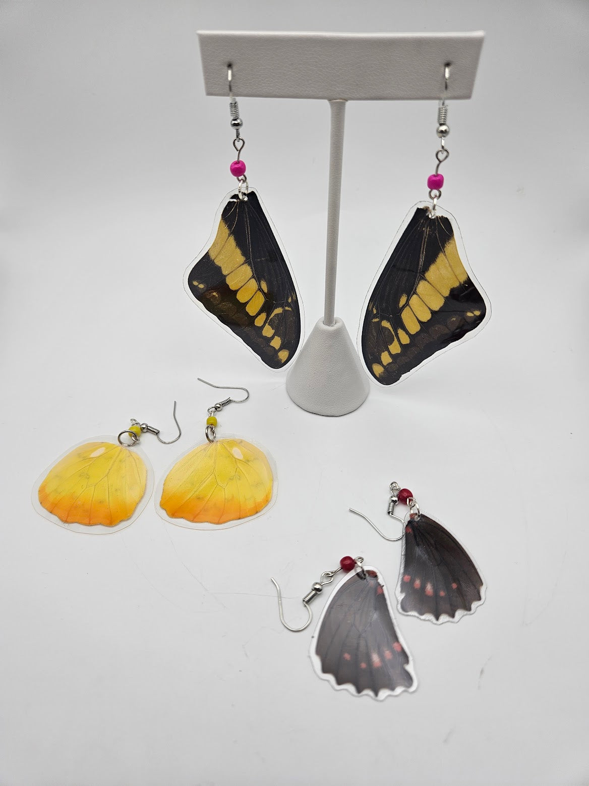 Three pairs of stunning Acrylic Butterfly Earrings are showcased on a white display. The mounted pair highlights black and yellow wings, while the two pairs below feature yellow and black wings adorned with pink spots, capturing the elegance of Butterfly Jewelry inspired by tranquil Butterfly Sanctuaries.