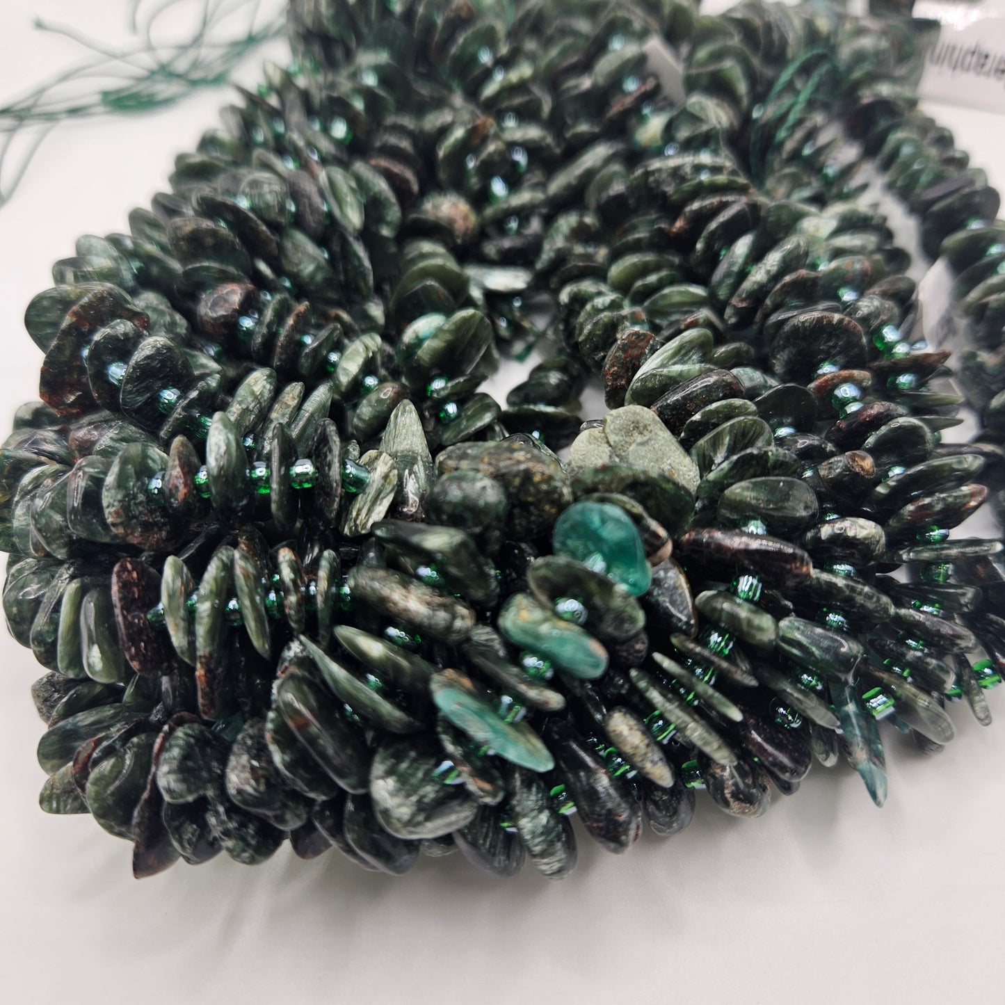Close-up image of multiple strands of Large Seraphinite Chips Bead Strand, featuring dark green, irregularly shaped polished beads. These seraphinite beads are shiny and vary in size, creating a textured appearance reminiscent of natural chips.