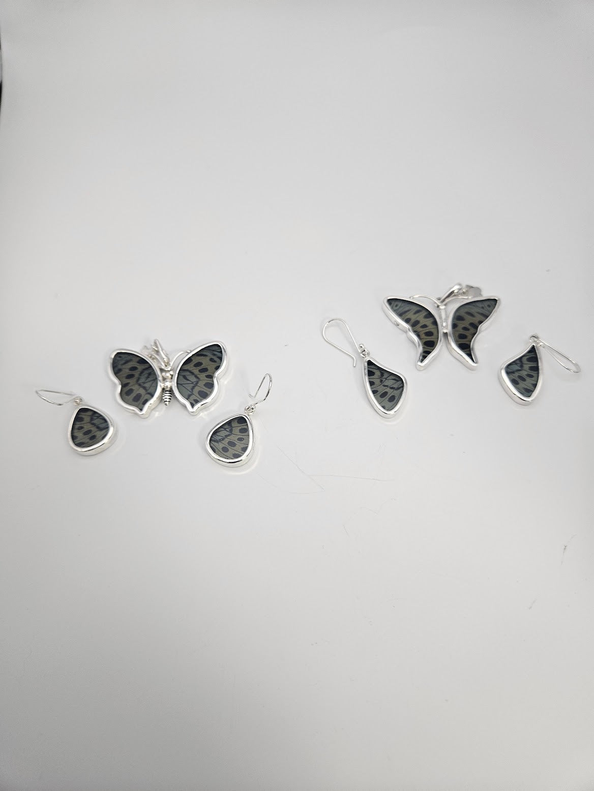 A display of two Sterling Silver Butterfly Small Sets on a white background captures the essence of a Butterfly Sanctuary. Each set includes three pieces: a brooch and a pair of earrings, adorned with black and gray patterned insets.