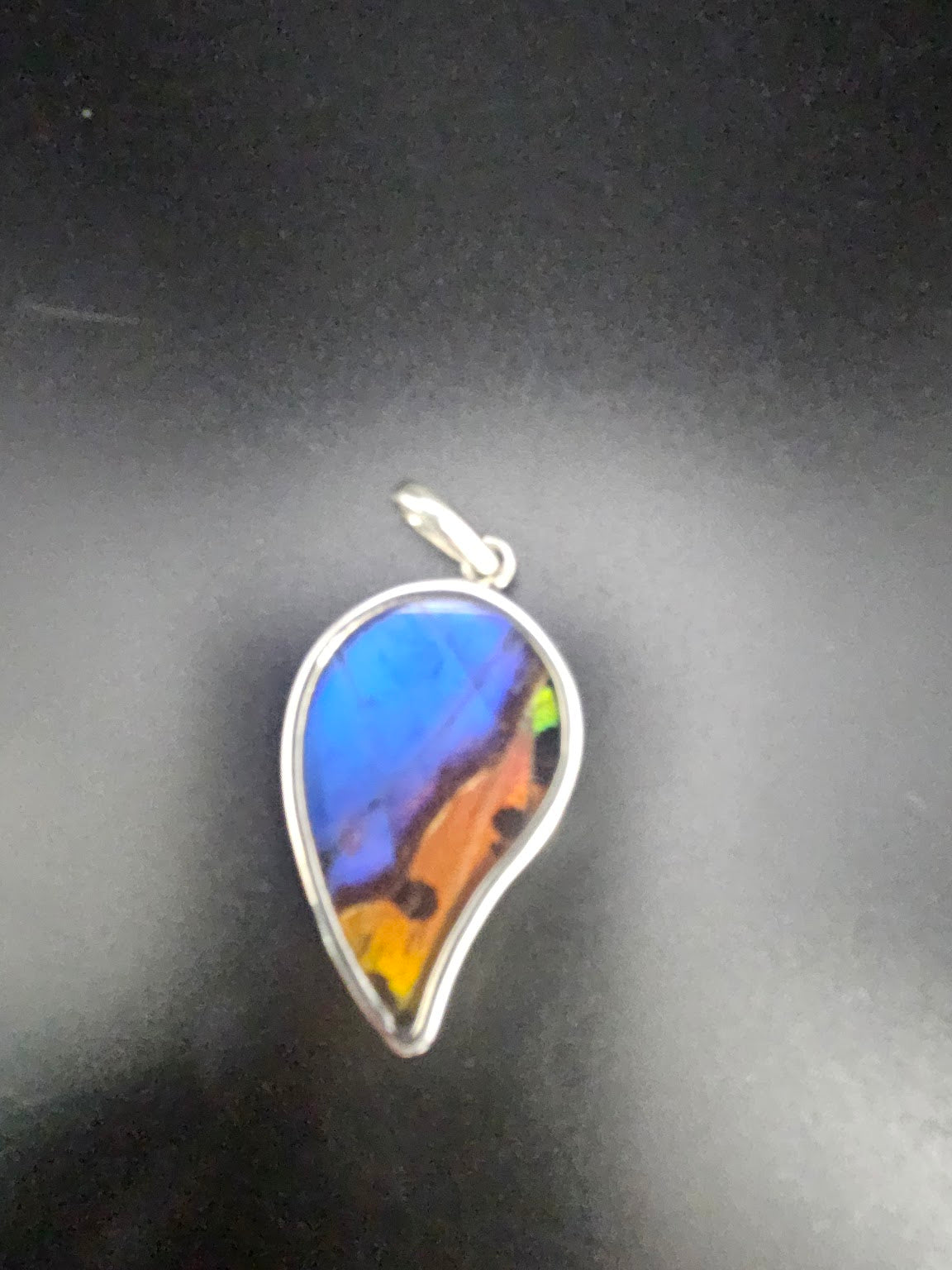 The Sterling Silver Medium Butterfly Wing Pendant features a teardrop design adorned with vibrant blue, orange, and black patterns reminiscent of butterfly jewelry, beautifully set in silver against a dark background.