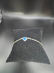 A sterling silver baby bracelet, featuring a heart-shaped blue charm and resembling a piece of jewelry art, is elegantly displayed on a black cushion.