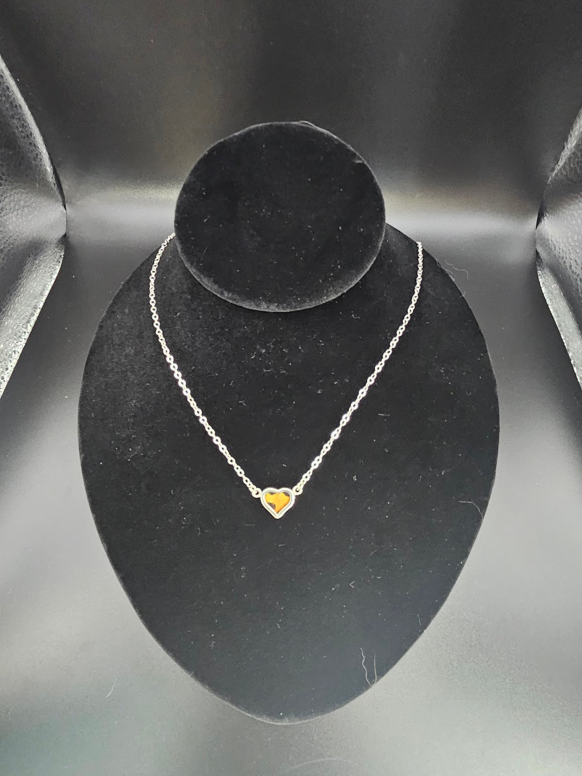 The Sterling Silver 1 Baby Necklace, featuring a small heart-shaped pendant, is beautifully displayed on a black velvet stand. The elegant gold-colored heart pendant offers a striking contrast against the silver chain, evoking the delicate beauty found in a butterfly sanctuary and resembling an artist's touch on nature's canvas.