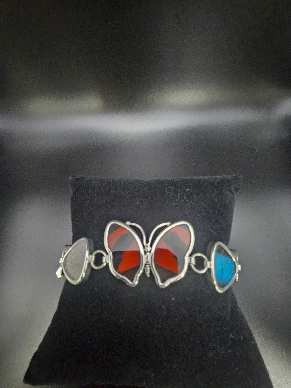 The Sterling Silver 5 Bead Butterfly Bracelet is a charming piece of jewelry featuring butterfly-shaped metal links with stained glass wings in shades of red, gray, and blue, reminiscent of a butterfly sanctuary. It is elegantly presented on a black cushion.