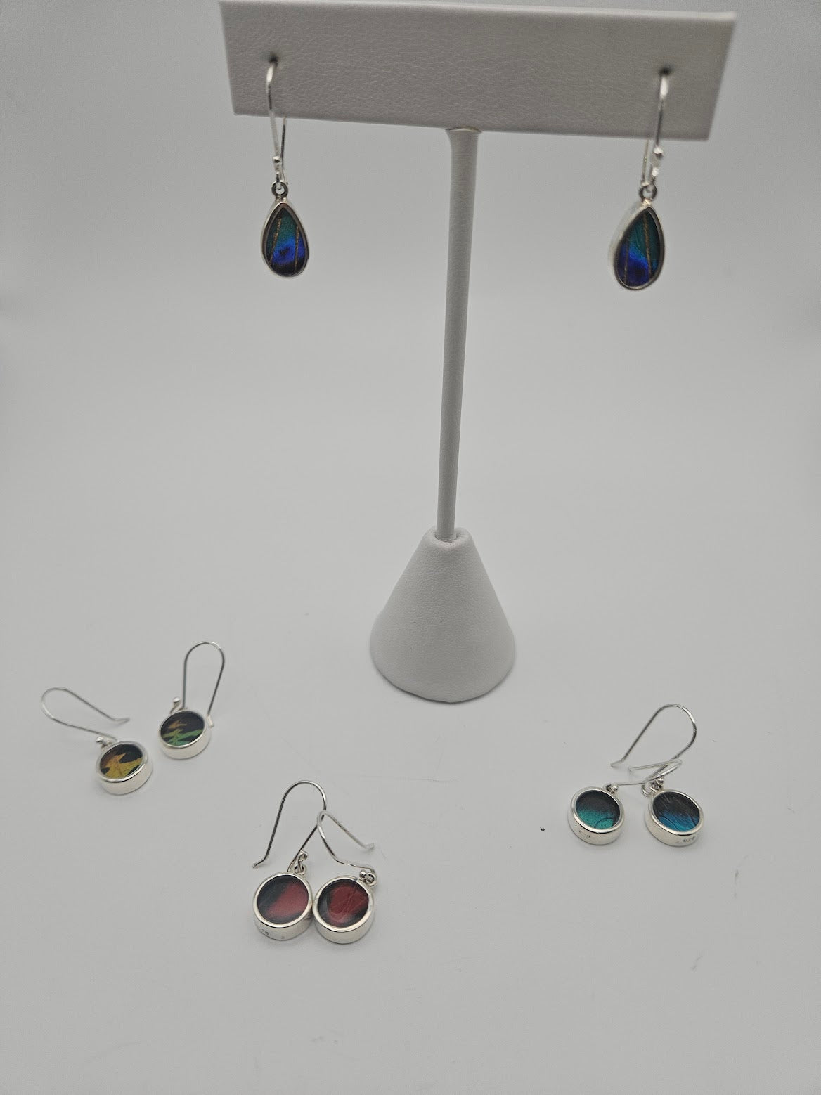 A display stand showcasing dangling earrings with teardrop-shaped iridescent stones takes center stage, reminiscent of treasures from a butterfly sanctuary. Encircling it are four pairs of Sterling Silver Butterfly Baby Earrings adorned with vibrant stones, all set against a plain white background, capturing the essence of Butterfly Jewelry.