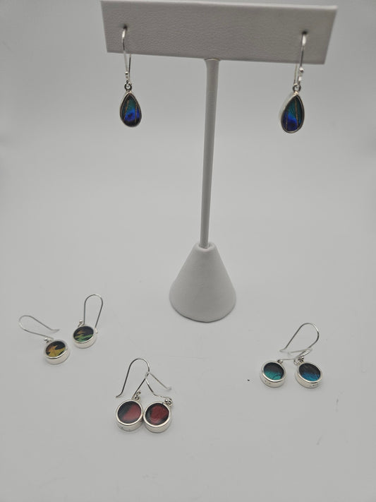 A display stand showcasing dangling earrings with teardrop-shaped iridescent stones takes center stage, reminiscent of treasures from a butterfly sanctuary. Encircling it are four pairs of Sterling Silver Butterfly Baby Earrings adorned with vibrant stones, all set against a plain white background, capturing the essence of Butterfly Jewelry.