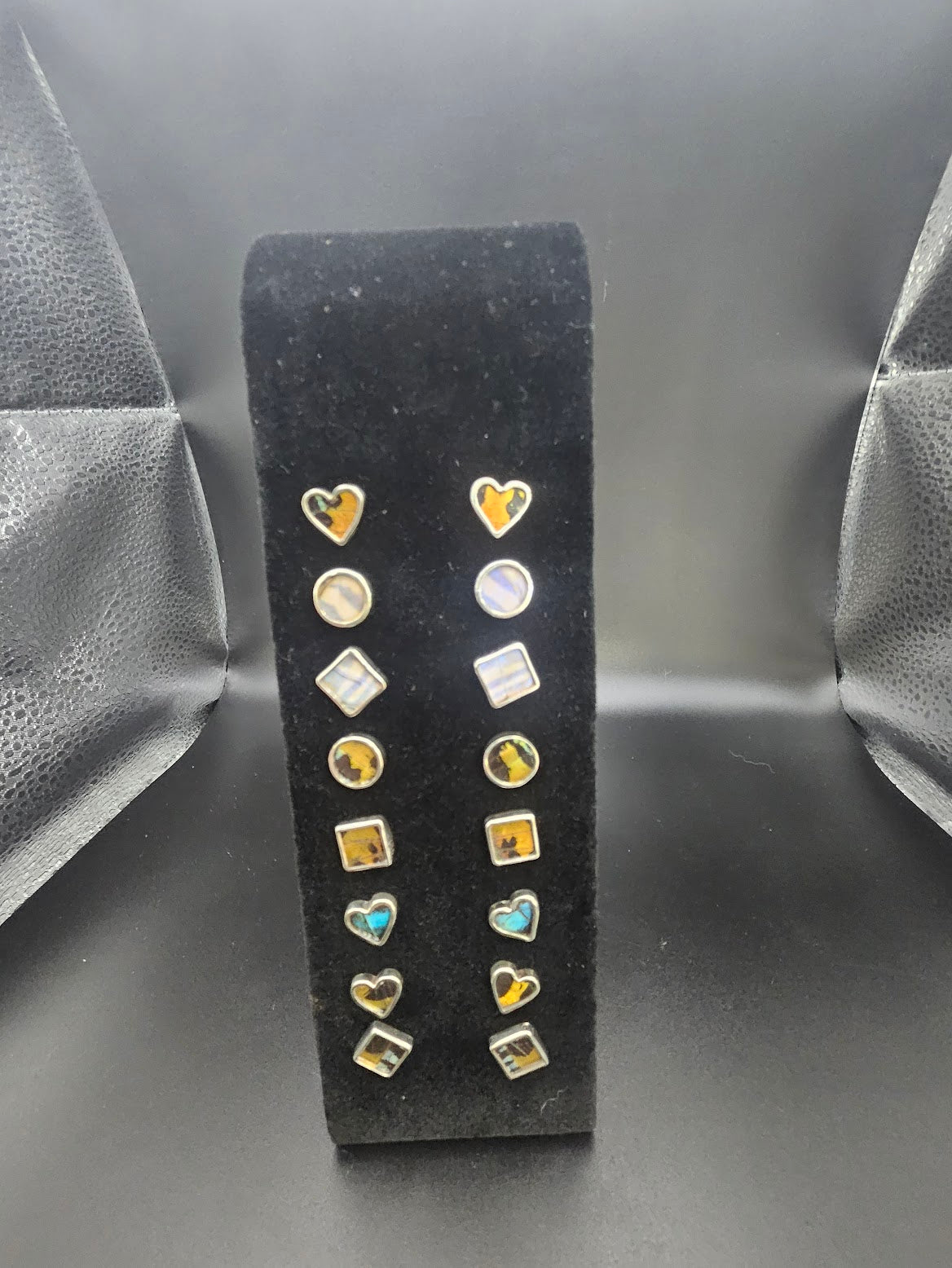 A black stand features a pair of Sterling Silver Butterfly Stud Earrings accompanied by heart and geometric designs in gold and silver tones, against a textured dark background reminiscent of a butterfly sanctuary.