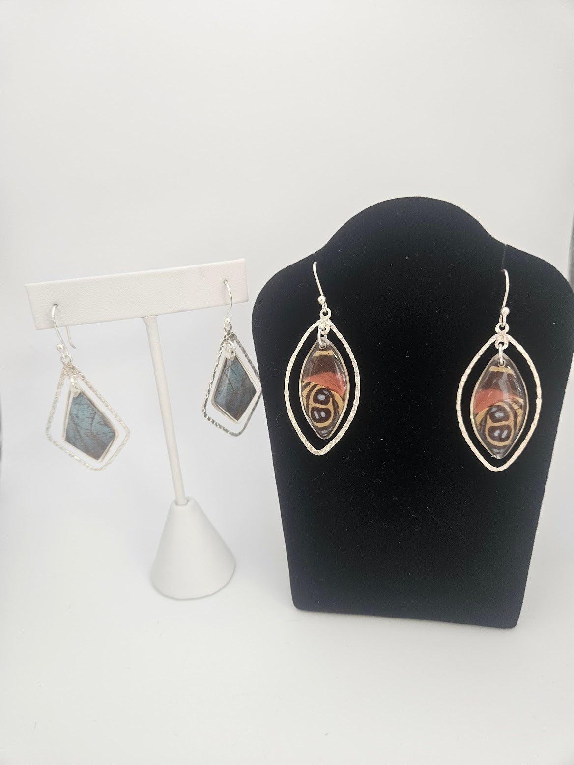 Displayed are two exquisite pairs of earrings: one featuring an abstract pattern on a black stand, and the other, the Sterling Silver Butterfly Ulises Earrings adorned with blue stone pendants reminiscent of delicate butterfly jewelry, showcased on a white stand against a plain background.