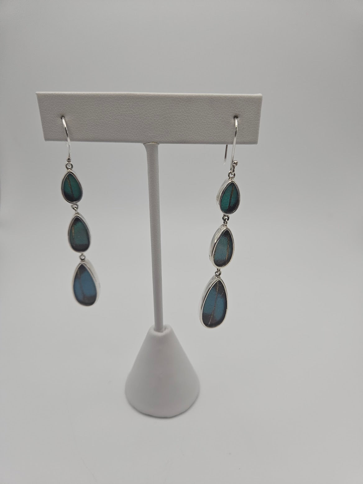 Displayed on a stand, the Sterling Silver Butterfly XL Tear Triple Earrings showcase three vertically aligned teardrop-shaped stones in a captivating deep blue-green hue. The plain, light gray background highlights their elegance, beckoning you to explore your second life's sanctuary of style.
