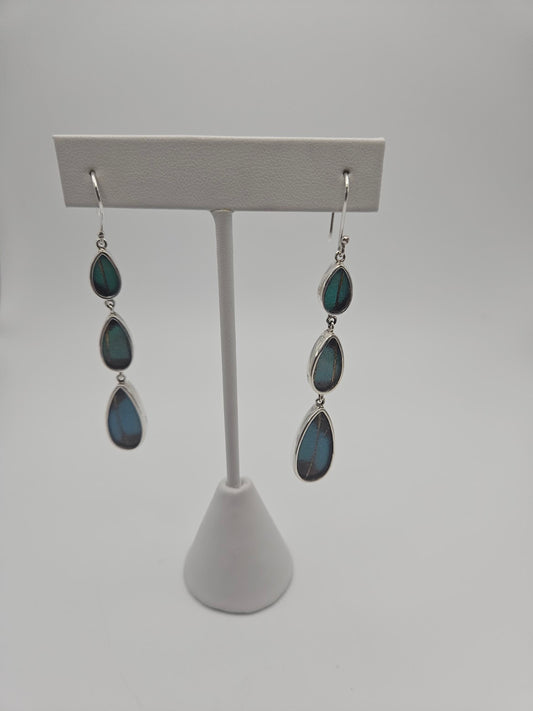 Displayed on a stand, the Sterling Silver Butterfly XL Tear Triple Earrings showcase three vertically aligned teardrop-shaped stones in a captivating deep blue-green hue. The plain, light gray background highlights their elegance, beckoning you to explore your second life's sanctuary of style.