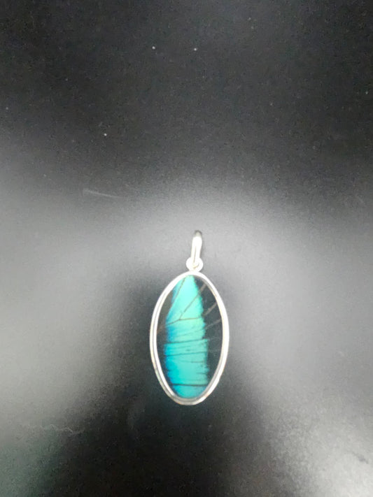 Introducing the Sterling Silver Medium Butterfly Wing Pendant, an oval-shaped accessory that showcases a captivating blue and green iridescent design, beautifully set in a silver frame against a dark backdrop.