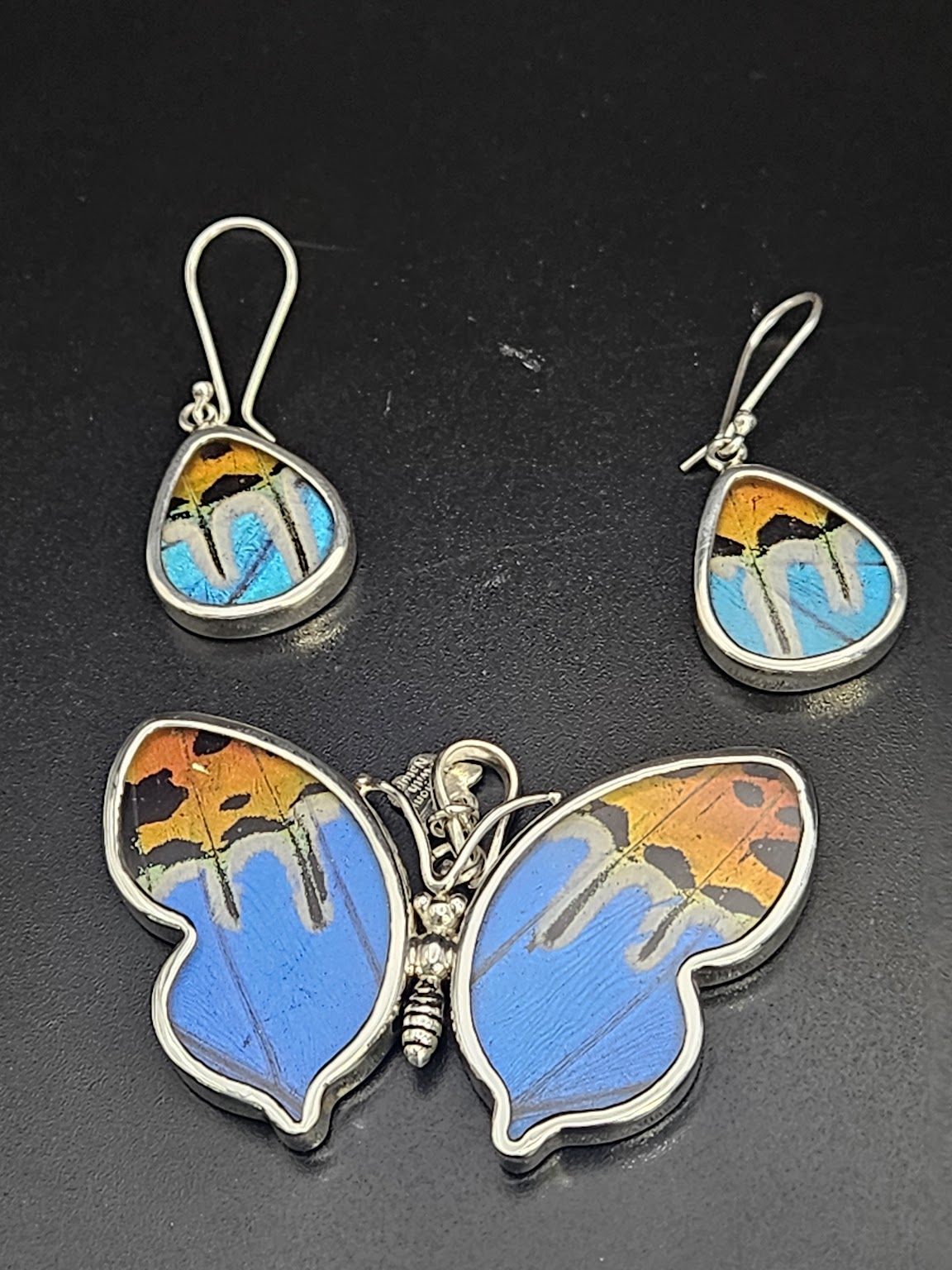 Explore the charm of the Sterling Silver Medium Butterfly and Wing Set, featuring exquisite teardrop-shaped earrings and a butterfly-shaped pendant. Each piece showcases a vibrant, multicolored design with shades of blue, orange, and black against a dark background, evoking the beauty of a butterfly sanctuary.