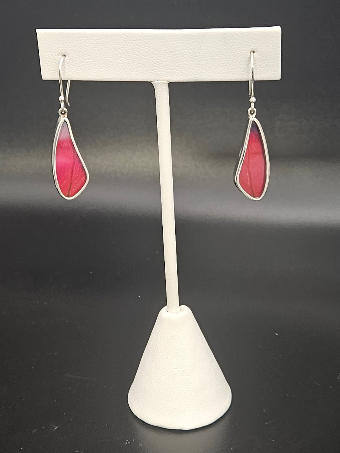 A pair of Sterling Silver Small Tear Wing Earrings with pink stones elegantly dangle like delicate treasures from the Butterfly Jewelry collection, hanging from a white earring stand against a dark background.