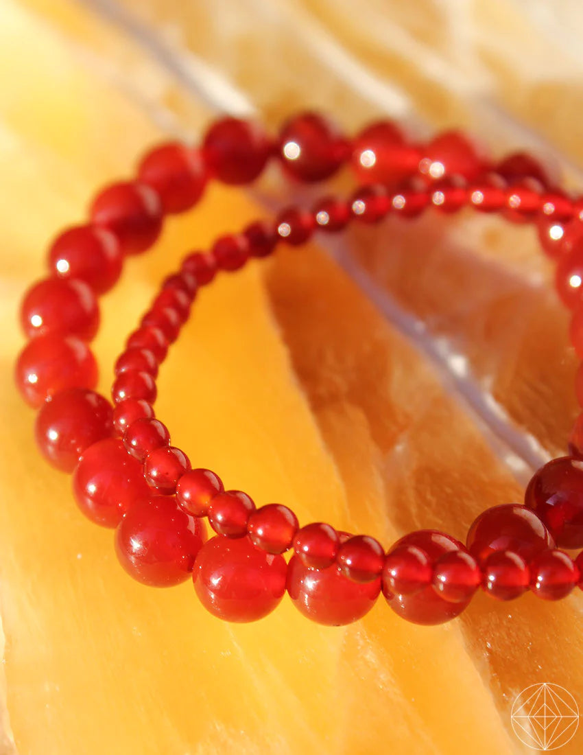 A close-up shot of two 8mm Carnelian bracelets resting on a textured, amber-colored surface. The larger beads on one bracelet contrast with the smaller beads on the other, creating a visually appealing arrangement that resonates with Root Chakra energy.