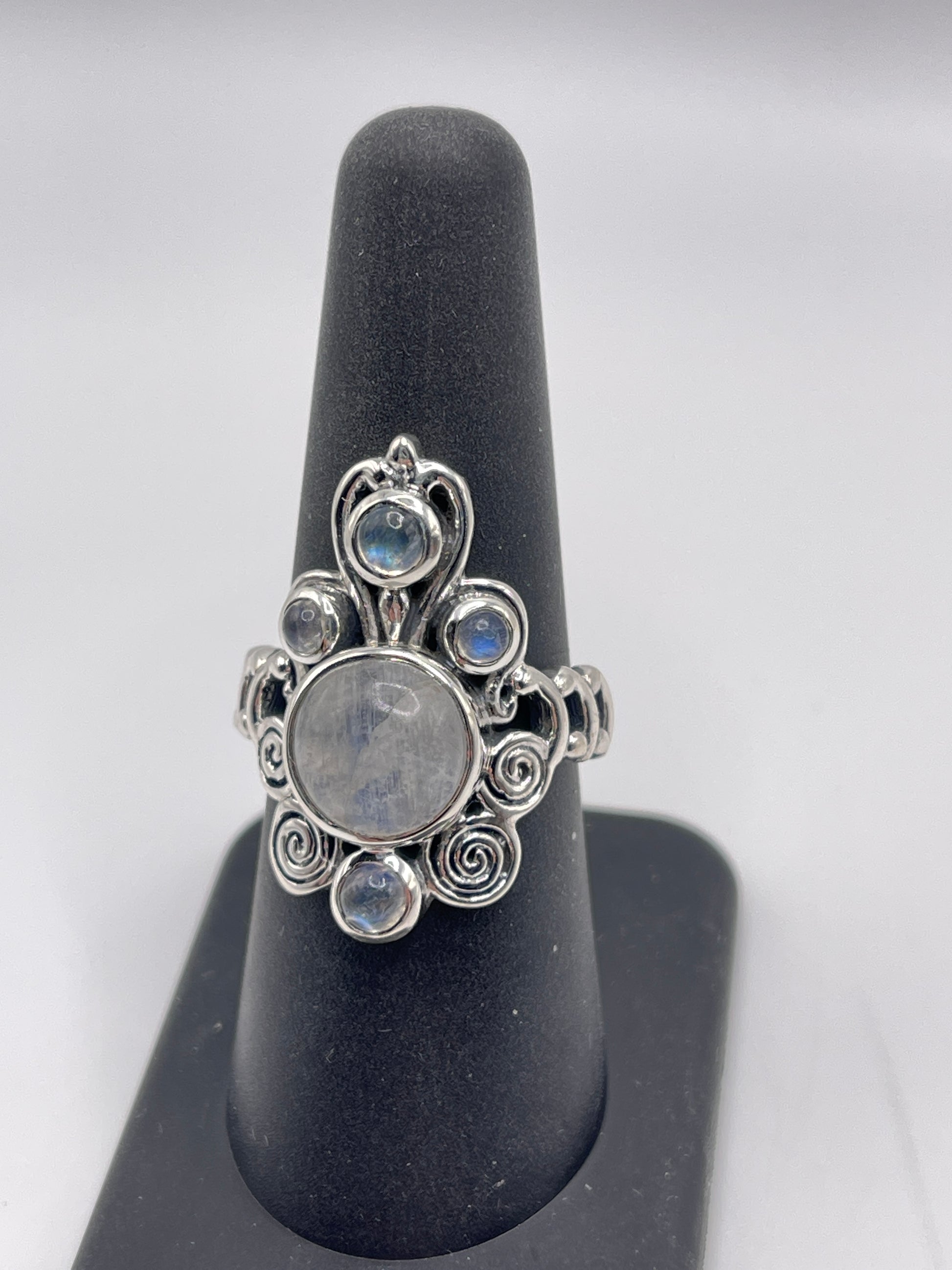 A "Moonstone Labradorite Swirl Statement Sterling Silver Ring Size 8" is elegantly showcased on a black stand, featuring a large round moonstone in the center, surrounded by smaller moonstones and intricate swirls. The background is a soft gray.