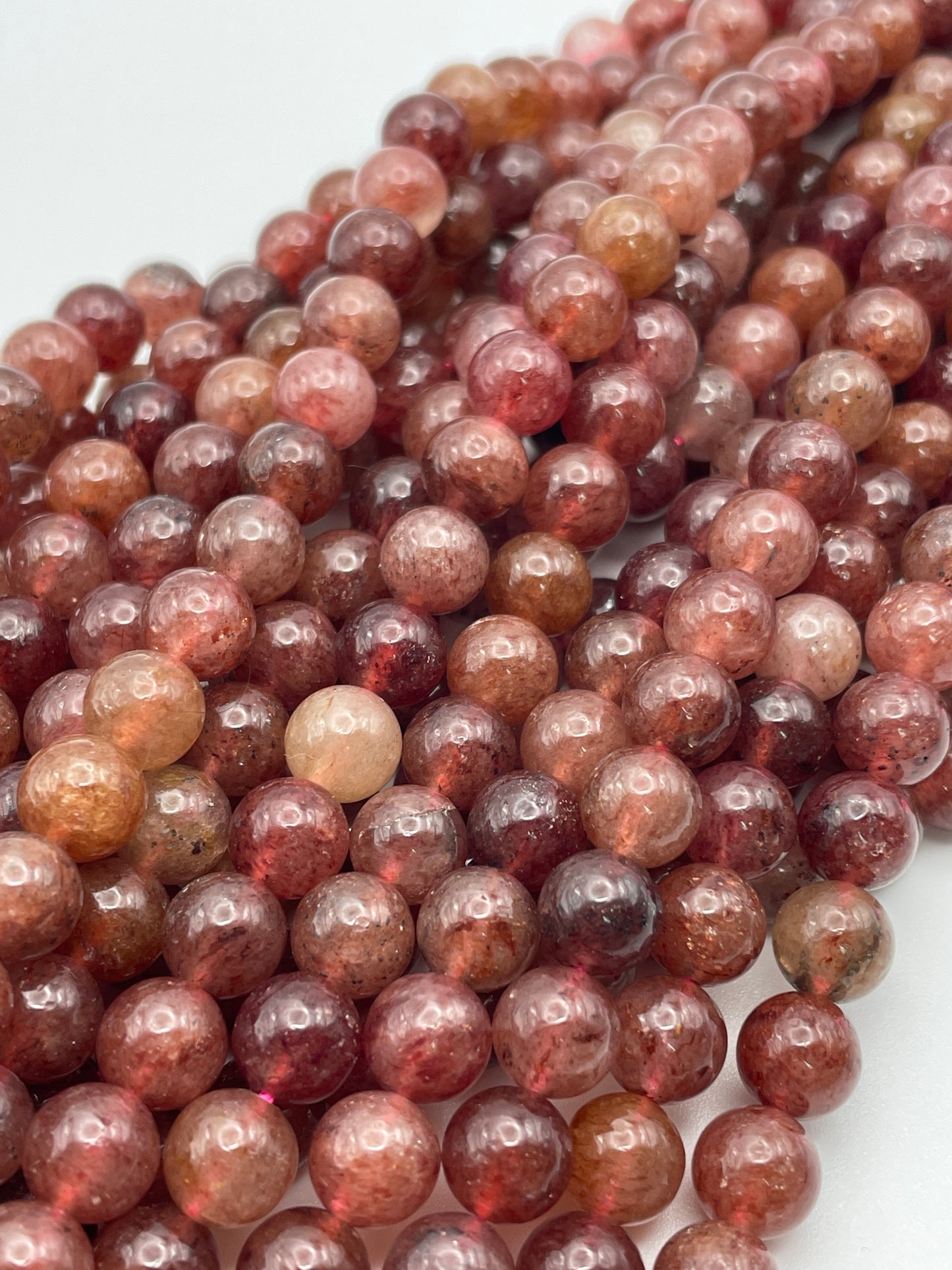 Close-up of several strands of 8mm Strawberry Quartz round beads, each polished to a reddish-brown hue with slight variations in shade, creating a textured surface. The glossy beads reflect light, giving them a shiny appearance.