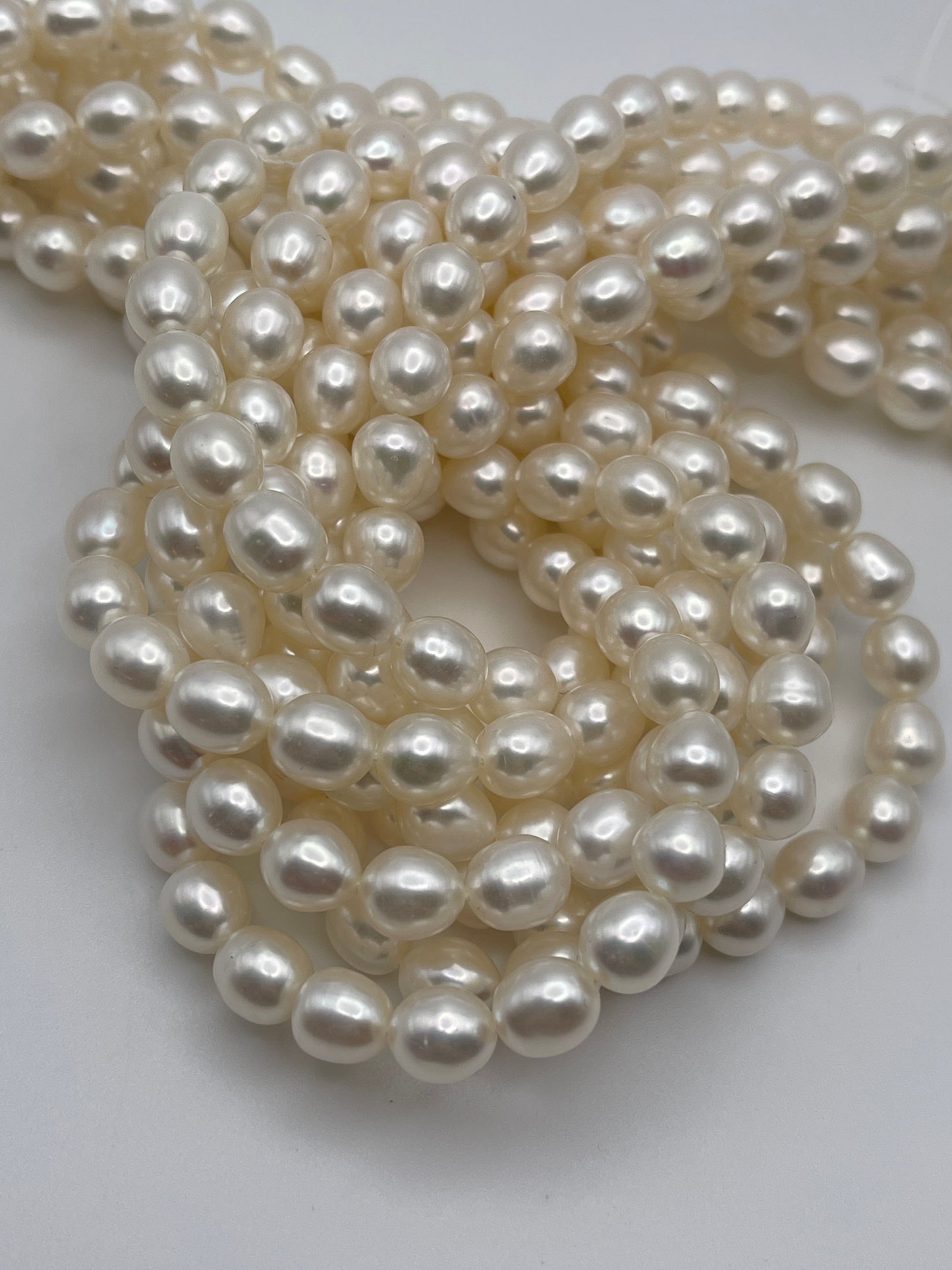A close-up image of several strands of 7.5-8mm white rice pearls piled together in loops, showcasing their smooth, shiny surfaces. The pearls have a slightly oval shape and a lustrous appearance against a plain white background.