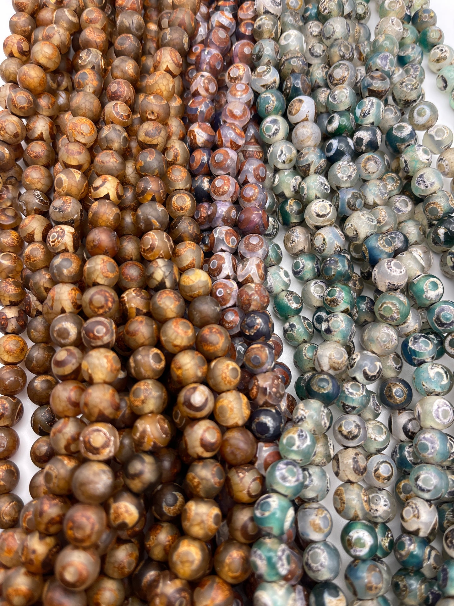 The 8mm Tibetan Agate Round IT strands feature multicolored beads arranged closely together, showcasing earthy tones such as browns, greens, and grays, with diverse natural patterns and textures.