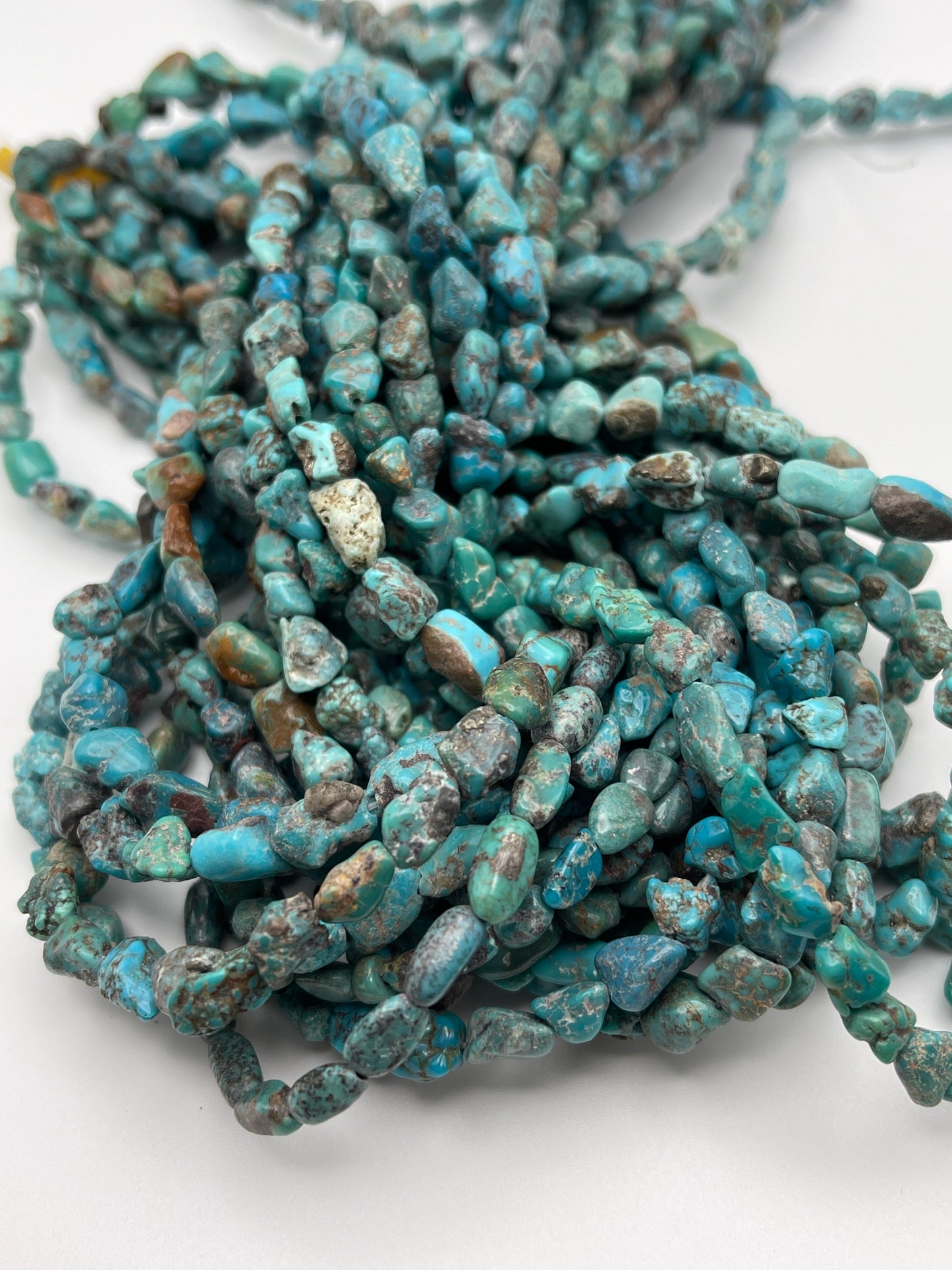 A close-up image of multiple 6-8mm Turquoise Chunk necklaces intertwined together. The beads are irregular in shape, displaying varying shades of blue and green with natural stone textures and patterns. The necklaces are set against a plain white background.
