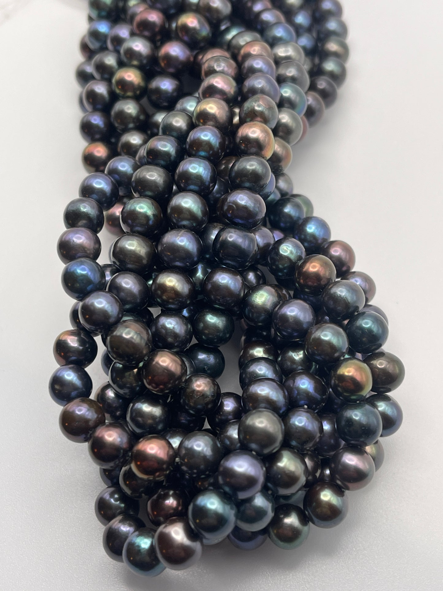 A close-up photograph of a tangled strand of 7-7.5 Peacock potato pearls, showcasing their iridescent, multicolored hues. The pearls have a lustrous finish and vary slightly in size, reflecting shades of green, blue, purple, and other dark tones.