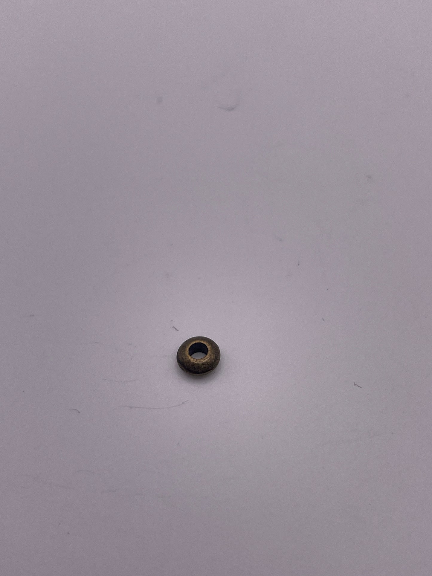 A Pewter Rondelle .35, a small, round bead with a hole in the center, sits on a white surface. The bead has a metallic finish and the lighting casts a faint shadow around it.