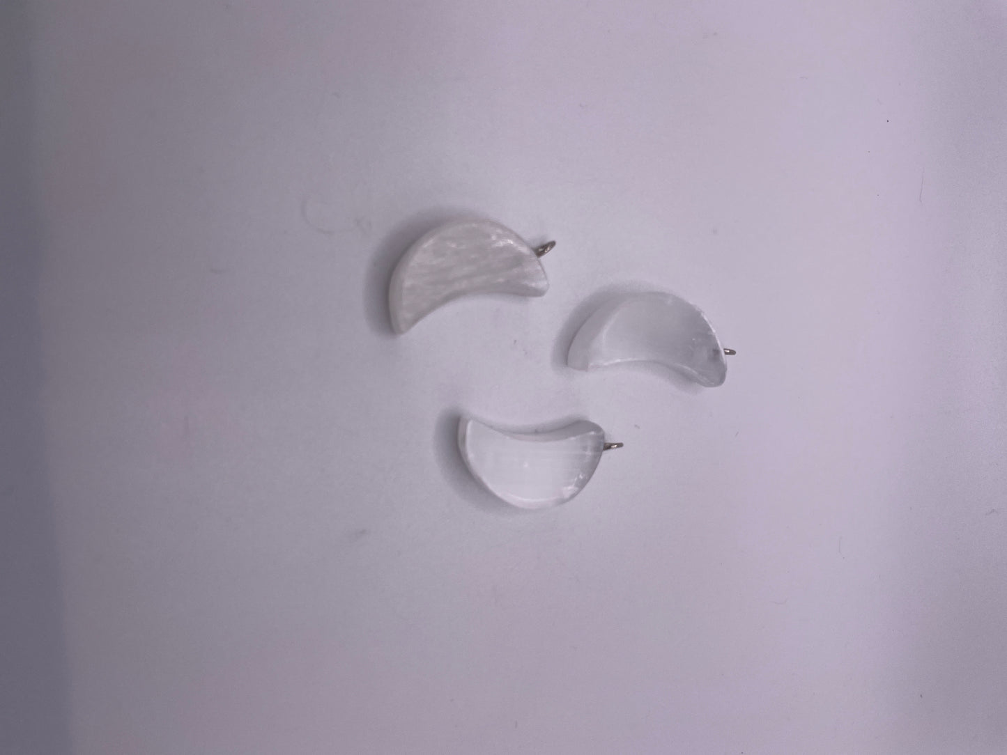 Three Selenite Moon Pendants, each measuring one inch and having a translucent crescent shape, are placed on a light gray surface. They appear to be small and gently curved.