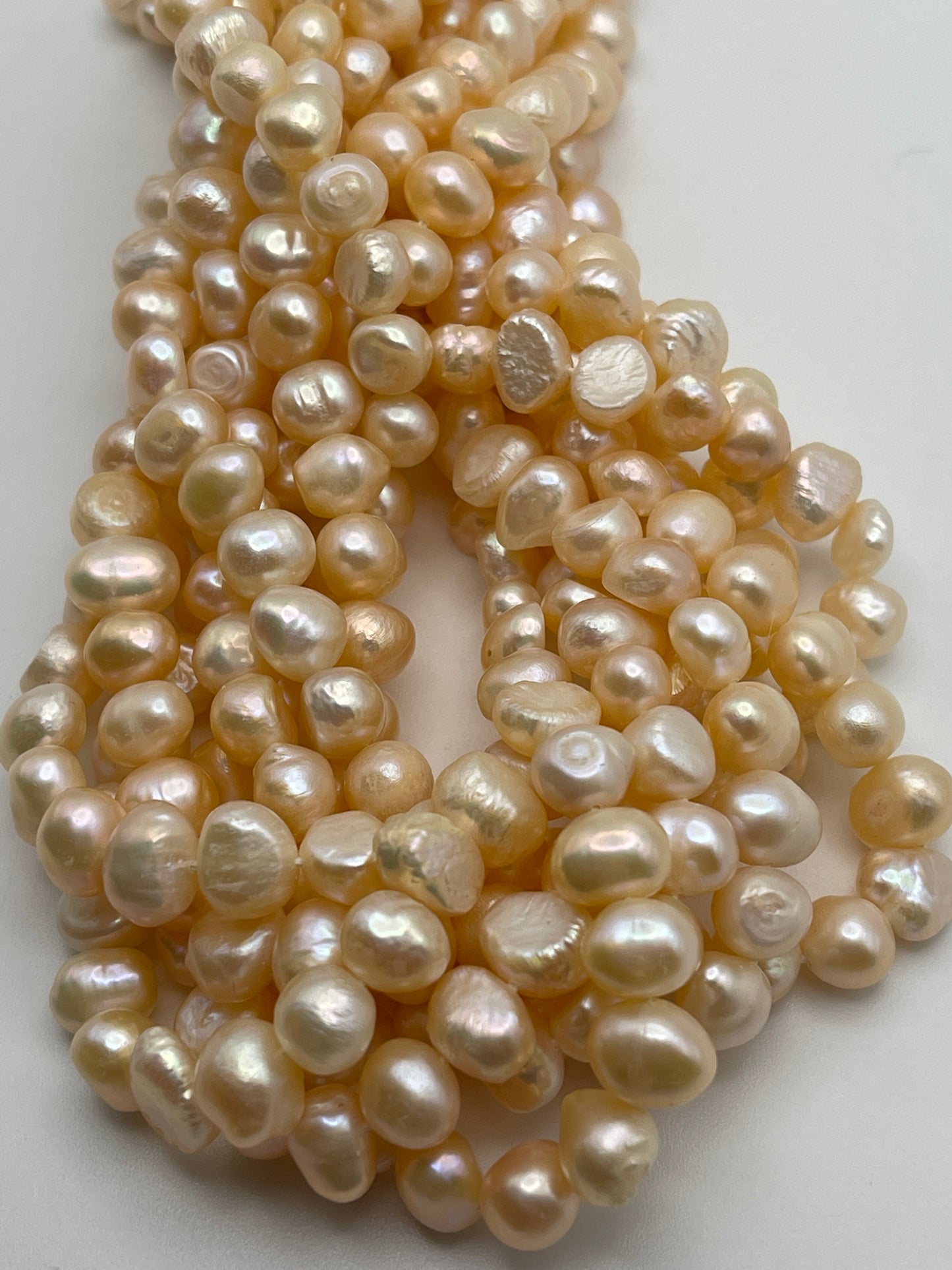A detailed close-up image displays multiple strands of lustrous, irregularly shaped, cream-colored 7-8mm peach flat pearls overlapping each other on a light background. The pearls have a shiny finish and exhibit a range of soft hues from pale yellow to soft pink.