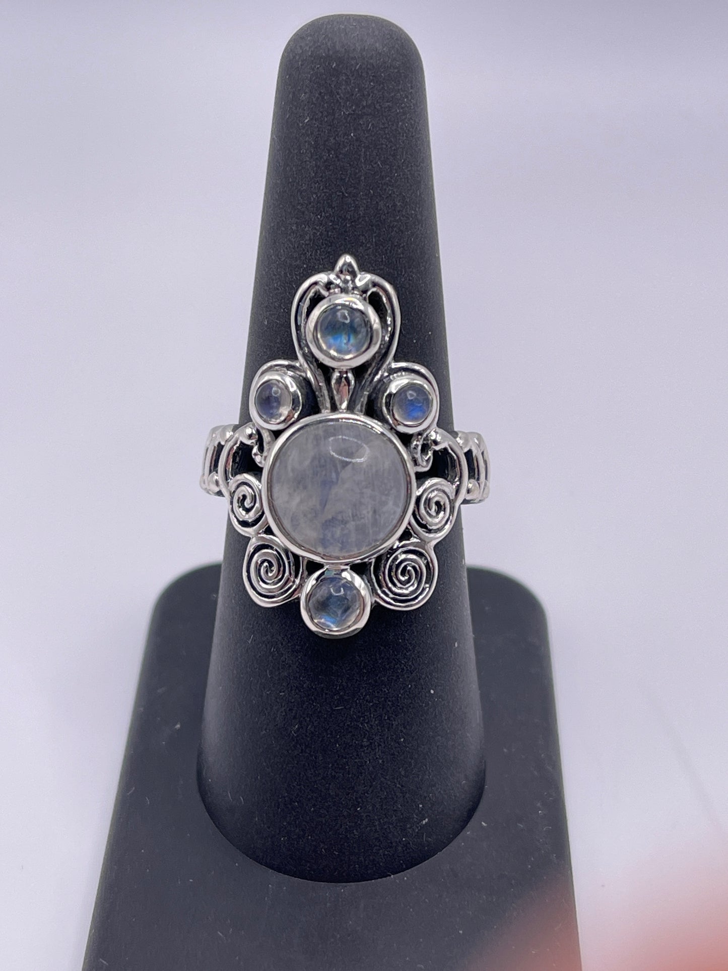 The Moonstone Labradorite Swirl Statement Sterling Silver Ring, Size 8, is elegantly showcased on a black ring stand. This decorative piece features intricate scrollwork and boasts a large, round central moonstone surrounded by smaller iridescent stones.