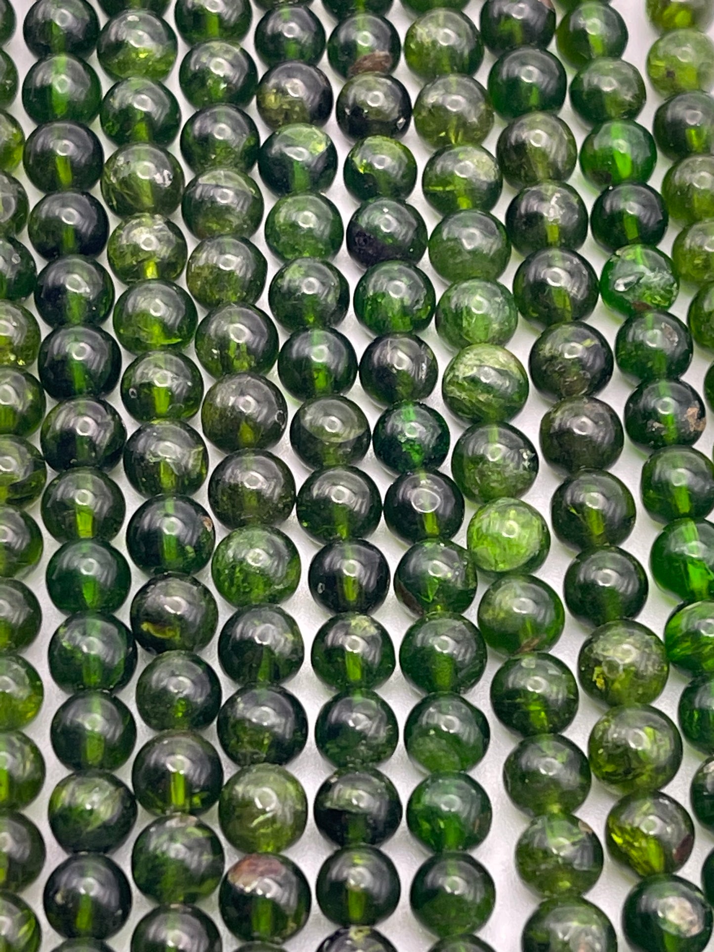 Rows of 5mm Chrome Diopside Rnd beads, polished to a high sheen, exhibit dark green hues interspersed with lighter green inclusions. The tightly arranged spherical beads reflect light off their smooth surfaces, creating a striking pattern.