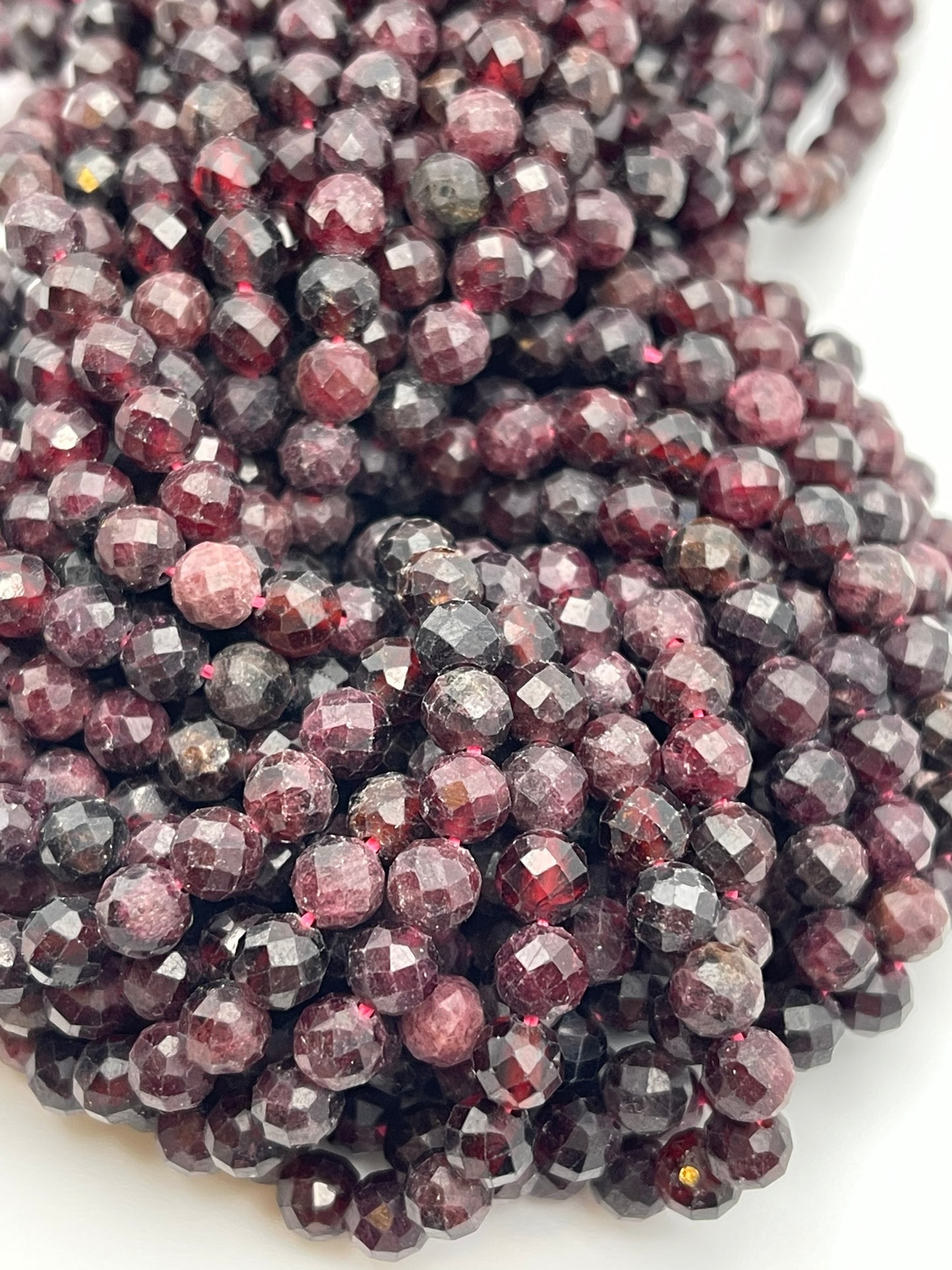 A close-up photo of numerous faceted, round-cut garnet beads from the 5mm Fac Garnet Rnd collection, arranged in dense, overlapping strands. The beads feature a glossy finish that reveals various shades of dark red and hints of blackish-red, reflecting light and highlighting their polished surfaces.