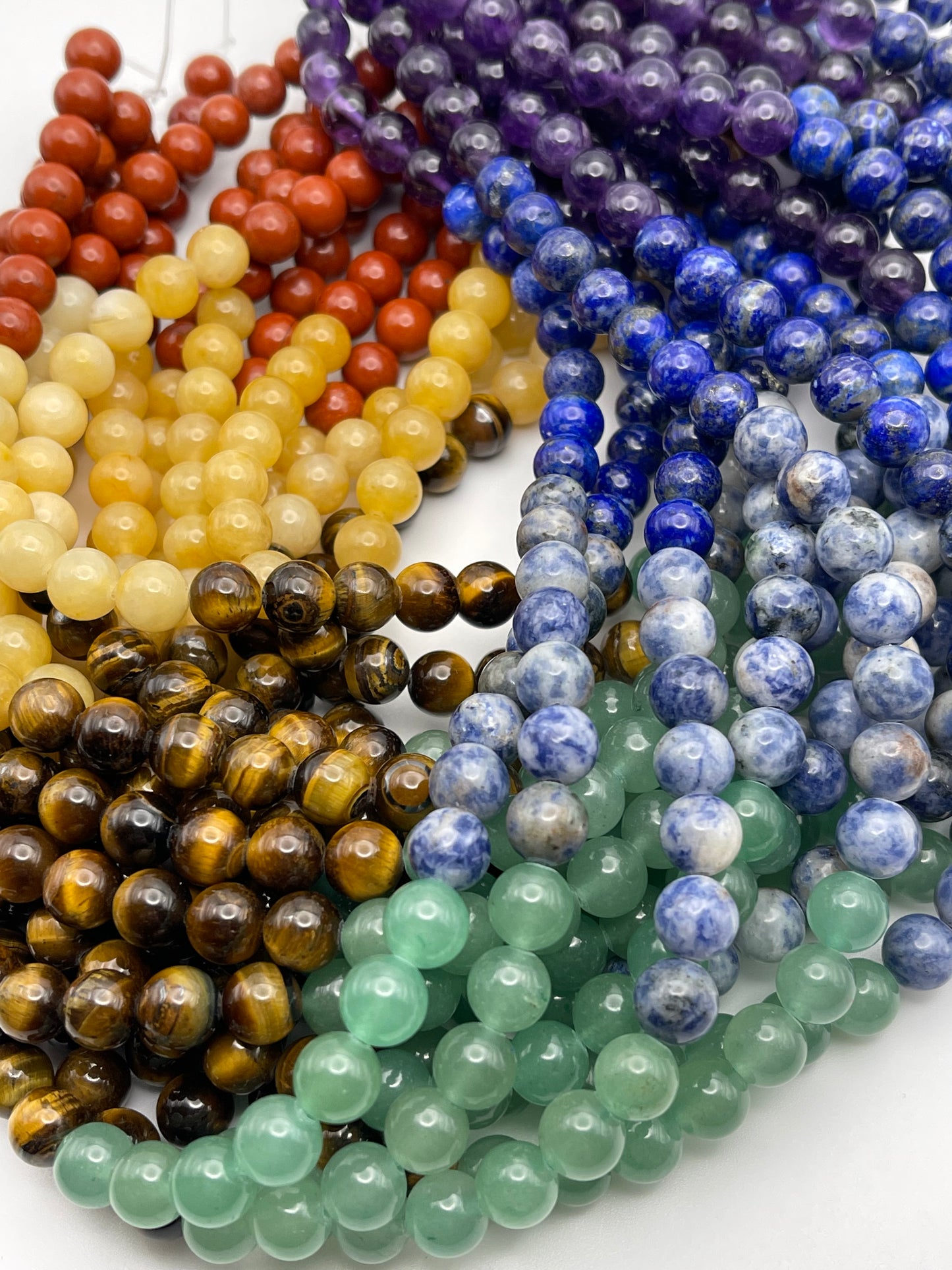 Strands of 8mm Round Chakra beads are laid out, showcasing an array of stones in vibrant colors. The collection includes red, amber, yellow, tiger's eye (dark brown with stripes), light green, blue, speckled blue and white, and deep purple stones. Each bead is shiny and polished to perfection.