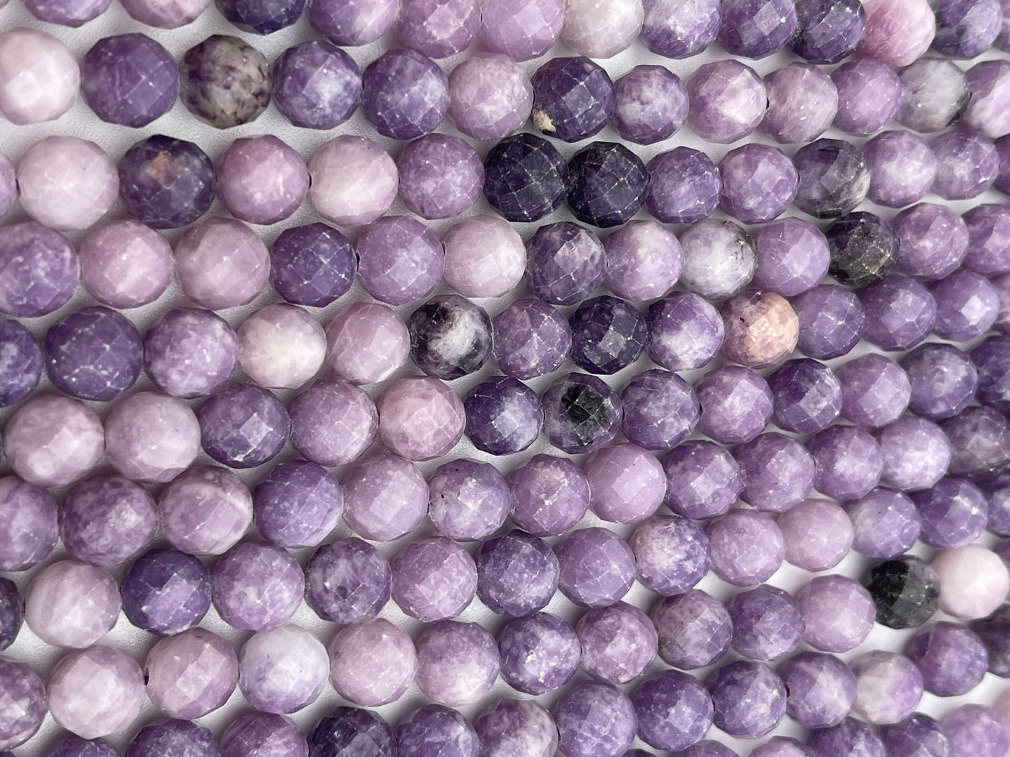 A close-up image of multiple rows of 8mm Fac Lepidolite Rnd beads placed side by side. The beads exhibit varying shades of purple with some marbled patterns, highlighting their texture and reflecting light.
